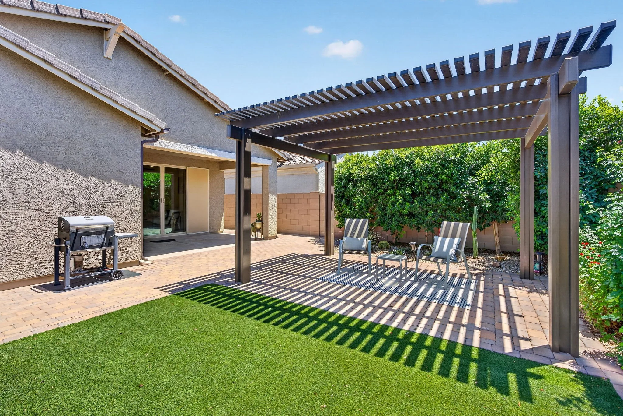 Backyard patio area with a wooden pergola casting shadows, two chairs, a grill, and a green lawn surrounded by trees and bushes.