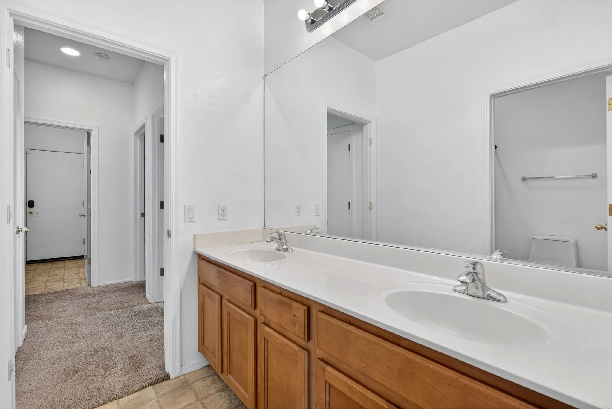 Bathroom with large mirror, white double sink vanity with wooden cabinet, white walls, towel rack, and a closed white door. Carpet and tile flooring.