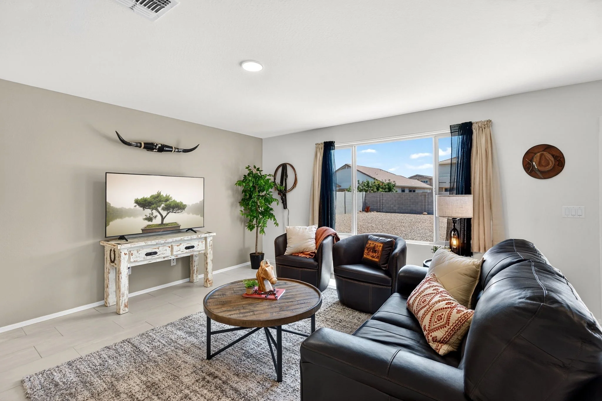 Living room with black leather sofa, beige armchair, round wooden coffee table, television on a distressed white stand, green potted plant, wall decorations including a hat and horn, large window with curtains, and beige carpet.
