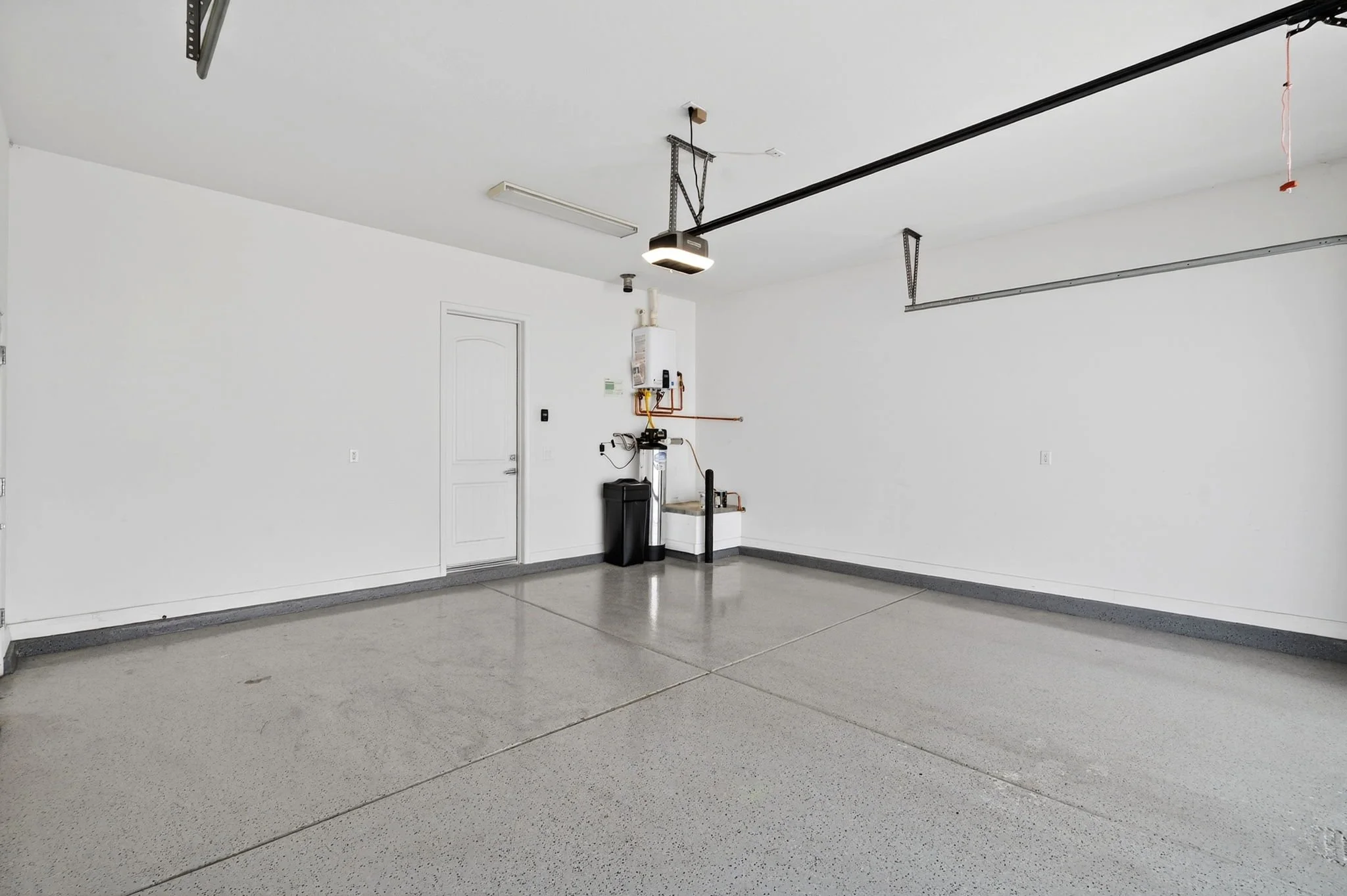 Empty garage with white walls, a closed white door, a garage door opener, and some tools and pipes on the wall.