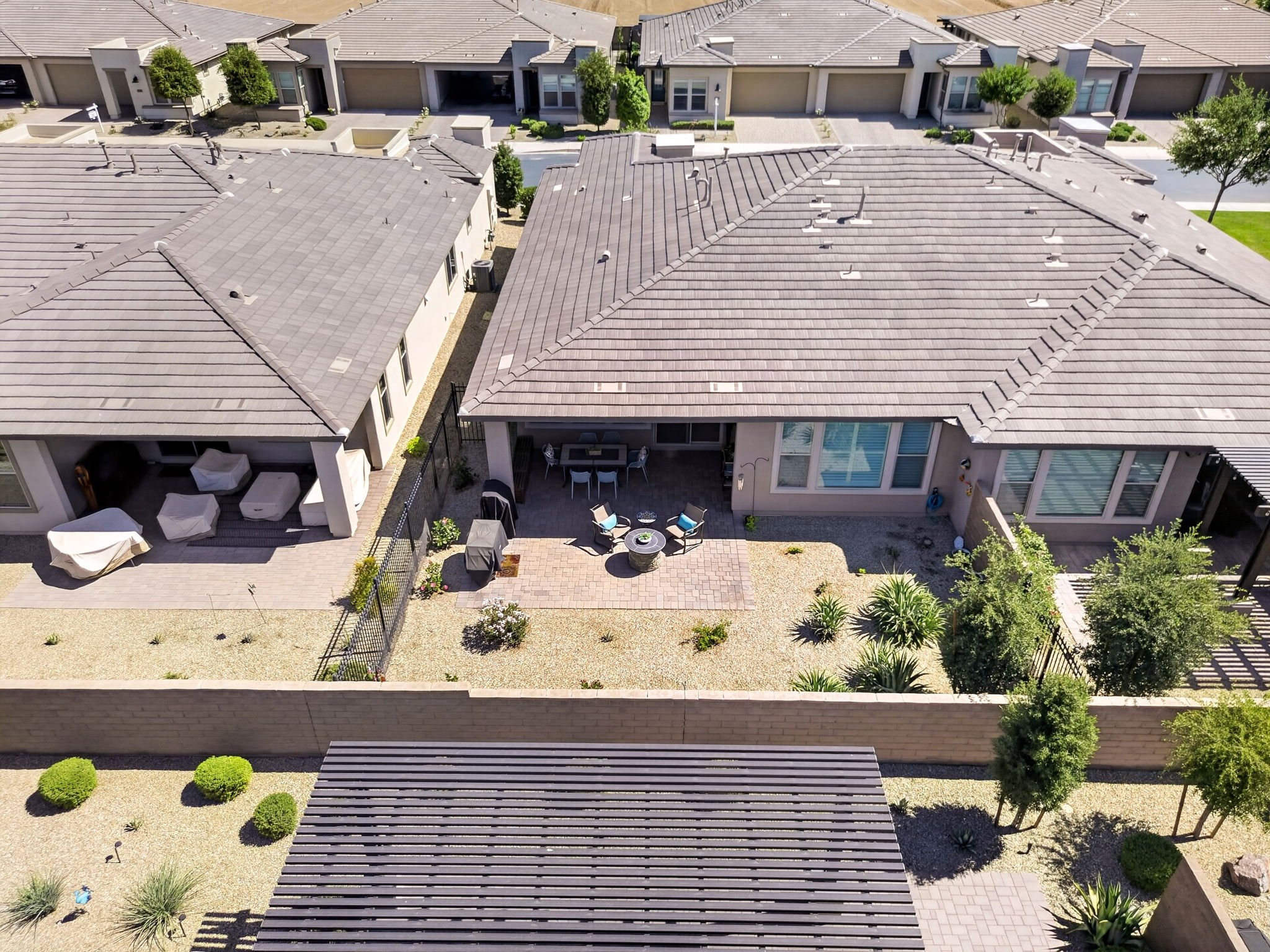 Aerial view of a residential backyard with a patio, outdoor furniture, and landscaped garden area, surrounded by neighboring houses with gray roofs.