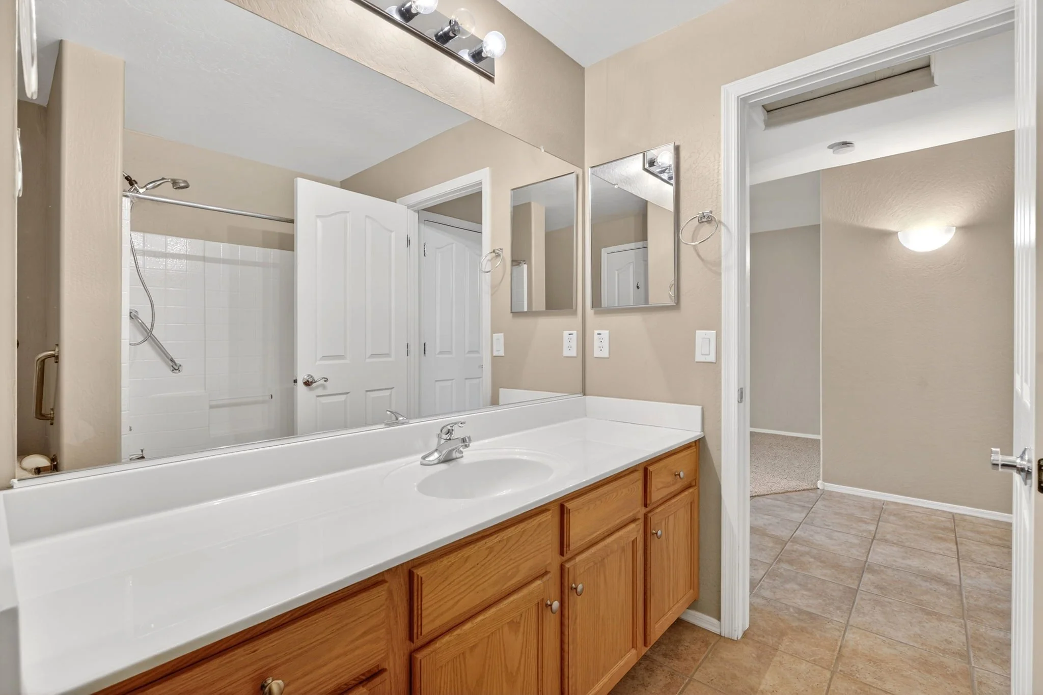 Bathroom with a large mirror above a white sink and wooden cabinets. A shower area with white tiles and a sliding shower door. Neutral beige walls and tile flooring. Overhead light fixtures and an open doorway leading to a carpeted room.