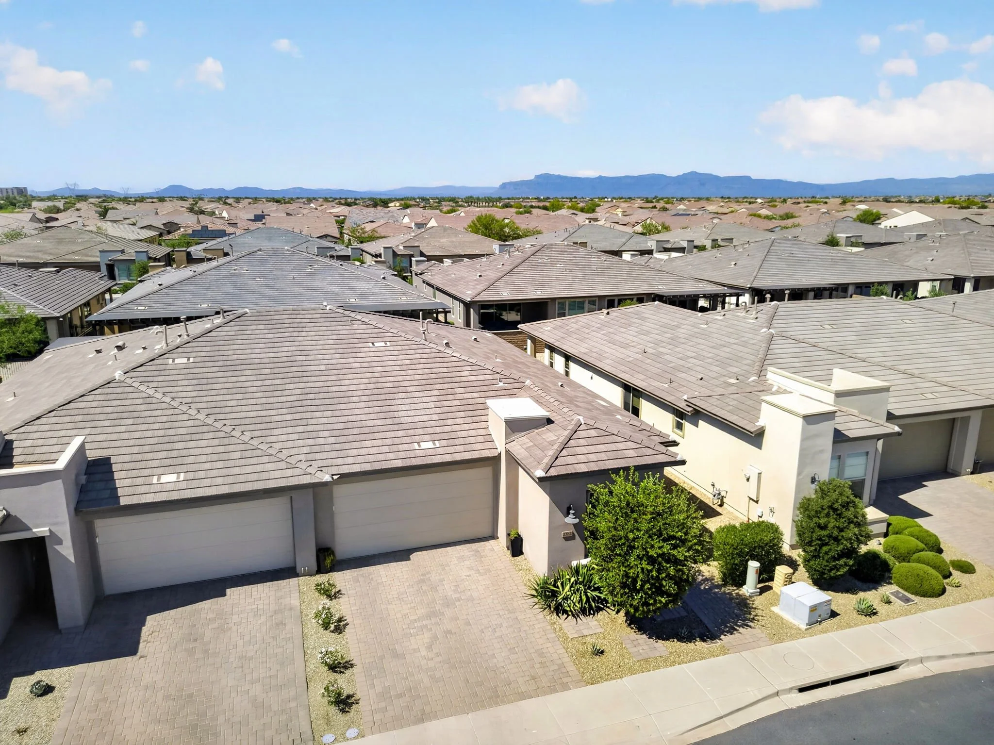 Aerial view of a suburban neighborhood with houses featuring grey and beige rooftops, small front yards with plants, and a paved driveway. In the background, distant mountains are visible under a blue sky with scattered clouds.