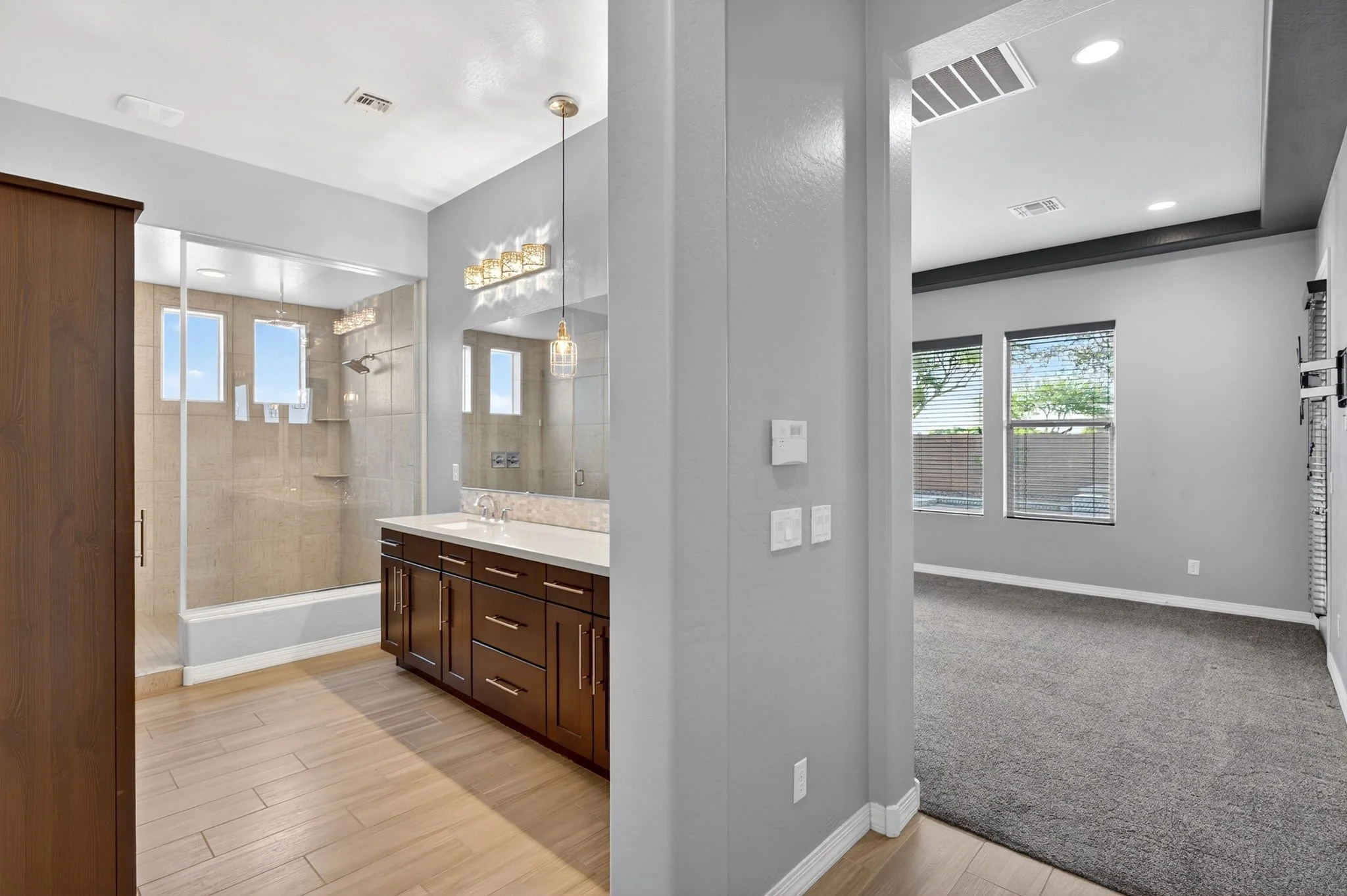 A view of a modern bathroom with a glass shower enclosure, beige tiles, a wooden cabinet vanity, and two pendant lights. Adjacent is a room with large windows, gray walls, and a carpeted floor.