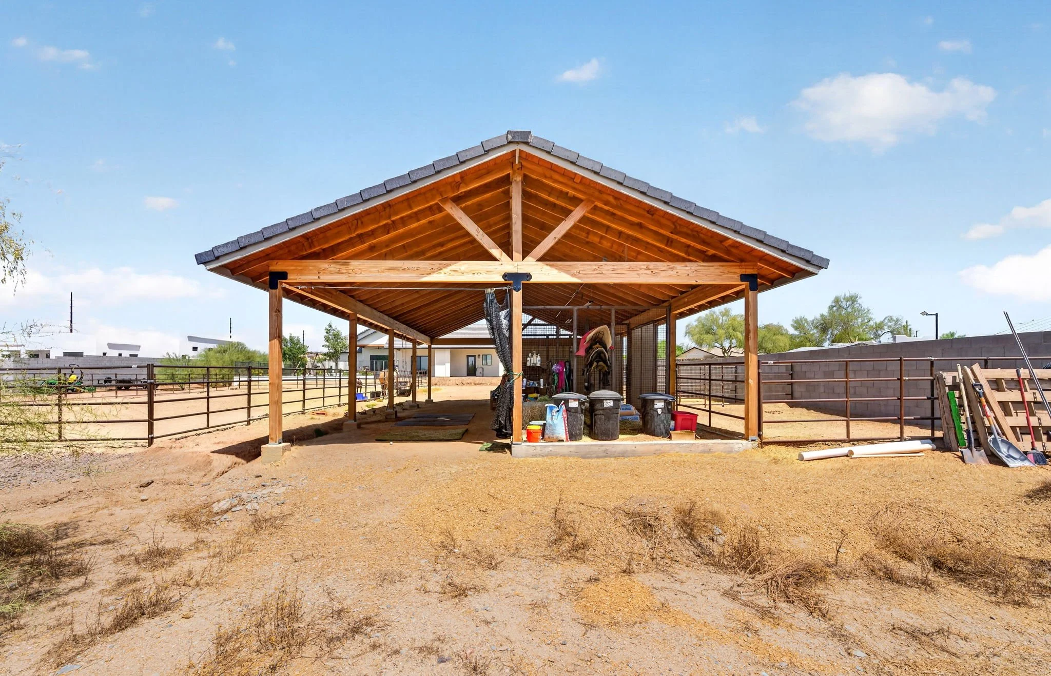 A wooden horse barn with a gabled roof, located in a yard with a dirt ground and fencing, containing various outdoor items and equipment, under a partly cloudy blue sky.