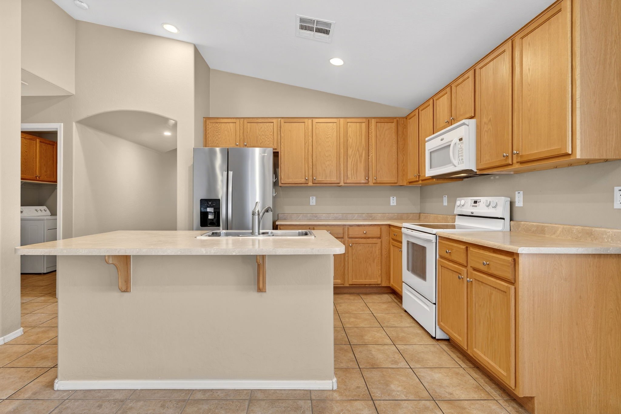 Kitchen with wooden cabinets, white appliances, beige countertops, and tile flooring, featuring a small island with a stainless steel sink.