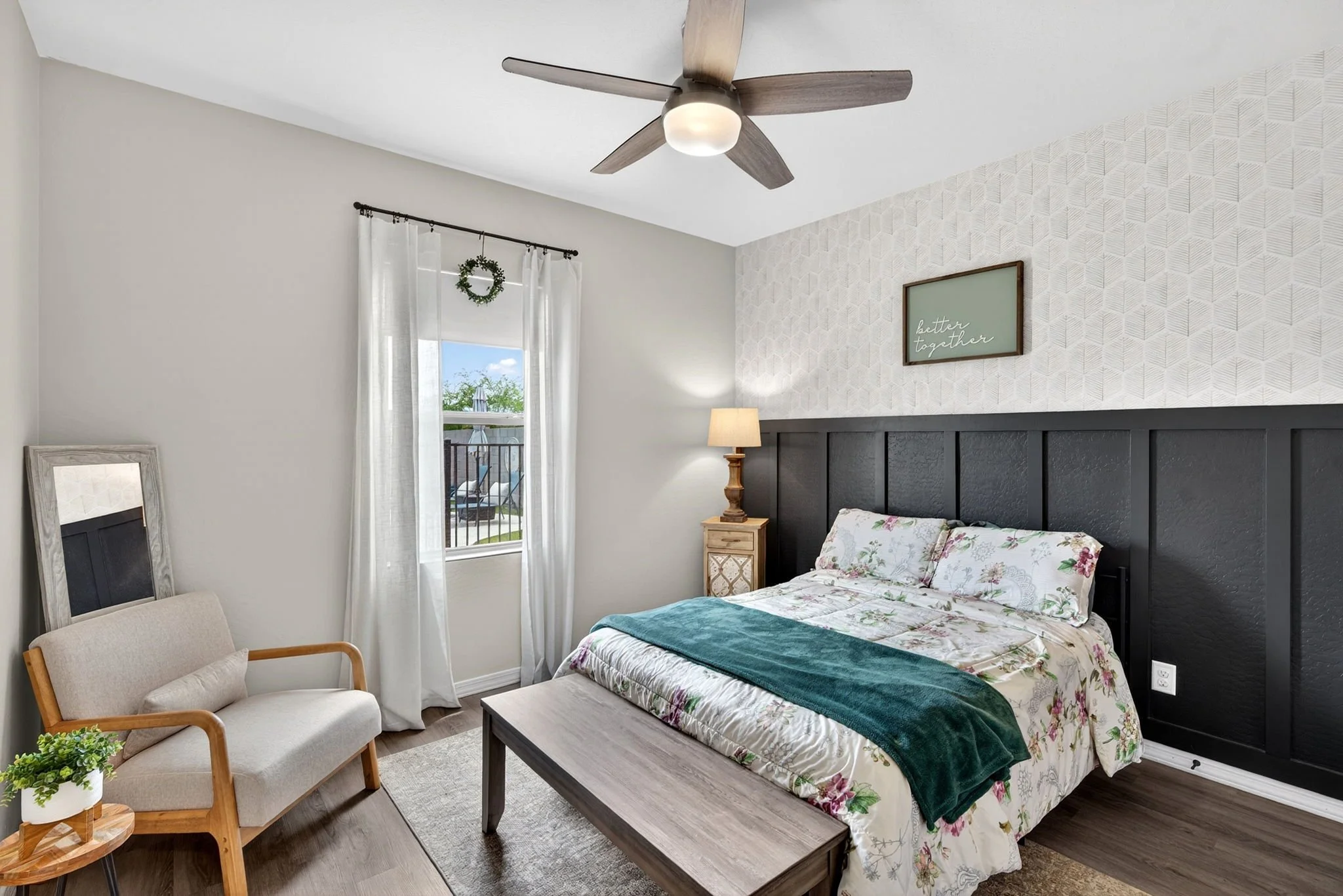 A cozy bedroom with a bed covered in a floral bedding set, a black headboard, and a green blanket. There's a wooden nightstand with a lamp, a picture frame with the phrase 'better together,' a window with white curtains and a wreath, a beige armchair