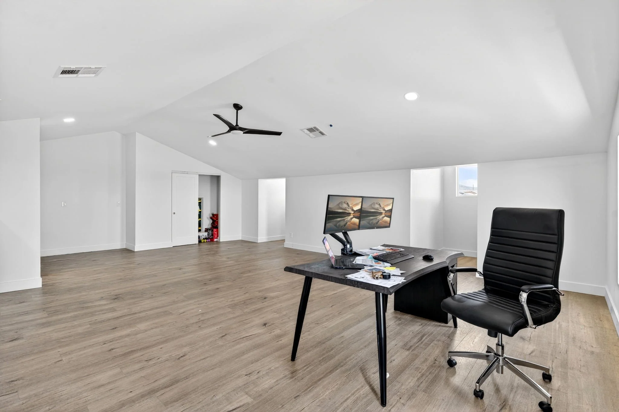 Empty office with a desk, computer, and black office chair on wood flooring, white walls, and a ceiling fan.