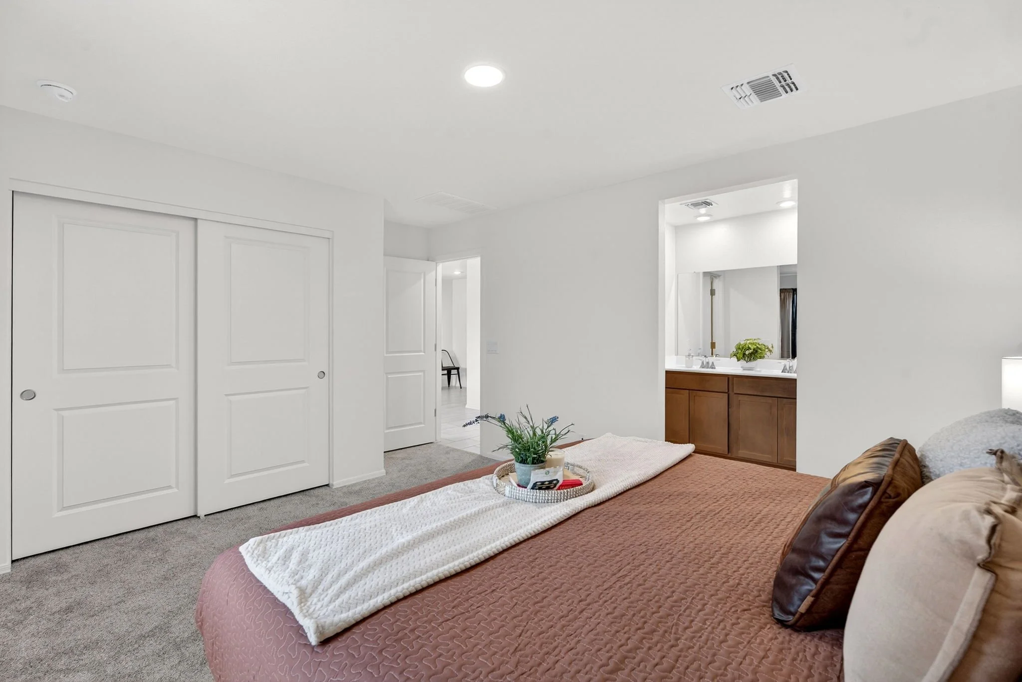 A bedroom with a bed, pillows, a tray with a remote and other items, a potted plant, and an open door revealing a bathroom with a vanity and mirror. The room has white walls, closet doors, and ceiling vents.