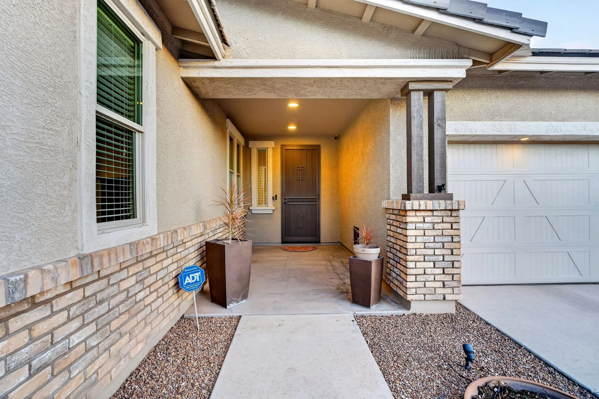 Front porch with potted plants, brick accents, and a closed garage door to the right.