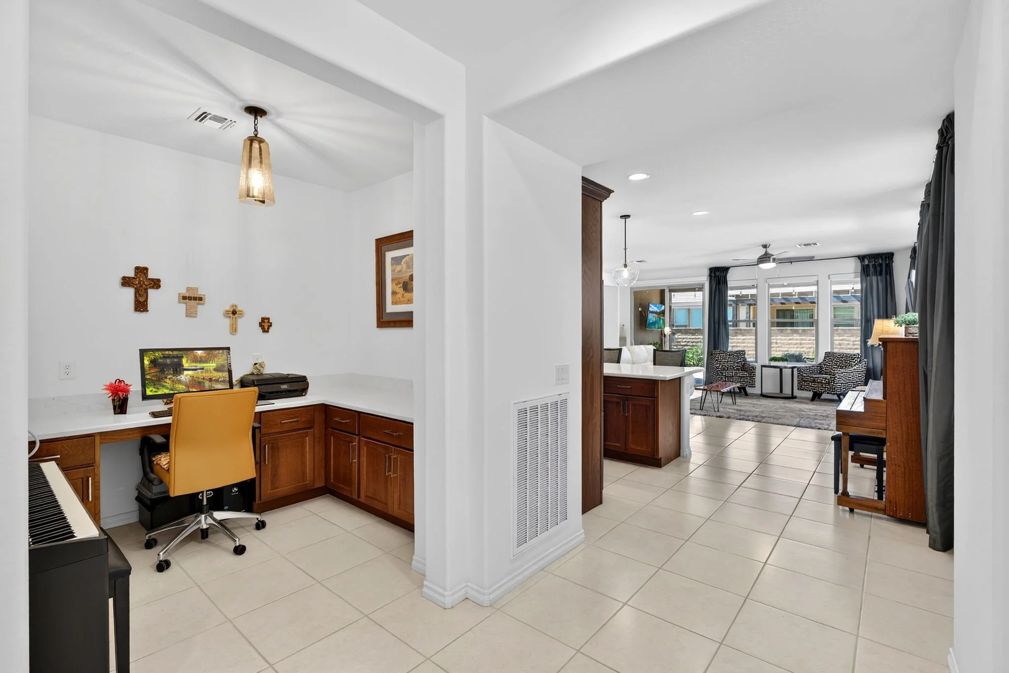 Living room with large windows, two black chairs, and a TV. Kitchen area with wooden cabinets and a countertop. White walls, tiled floor, and curtains on windows.