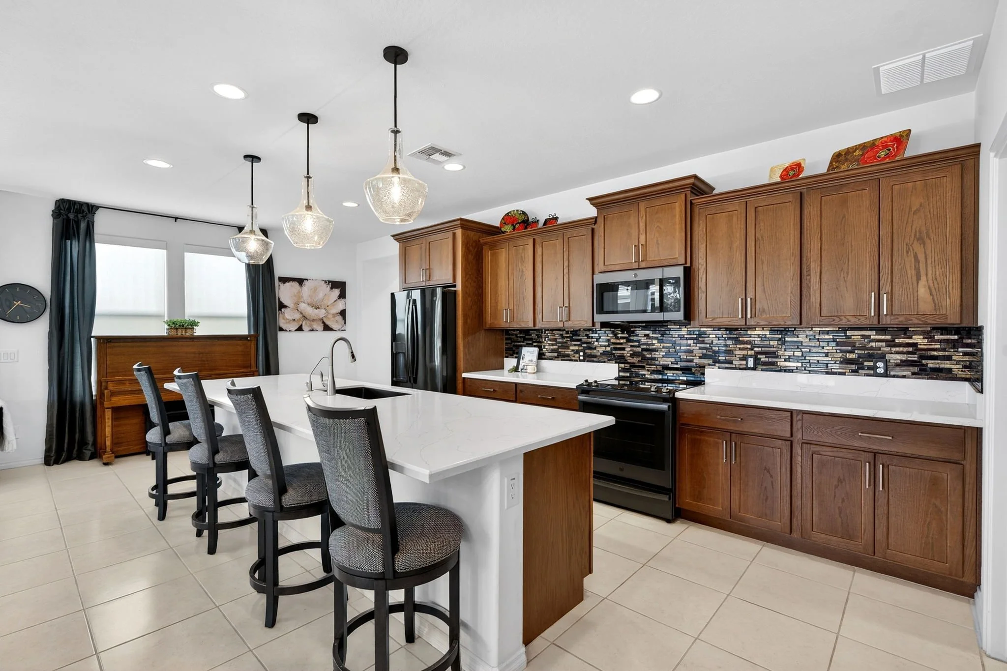 Modern kitchen with wood cabinets, a mosaic tile backsplash, stainless steel appliances, a white island with four black and gray chairs, pendant lights, and large windows with dark curtains.