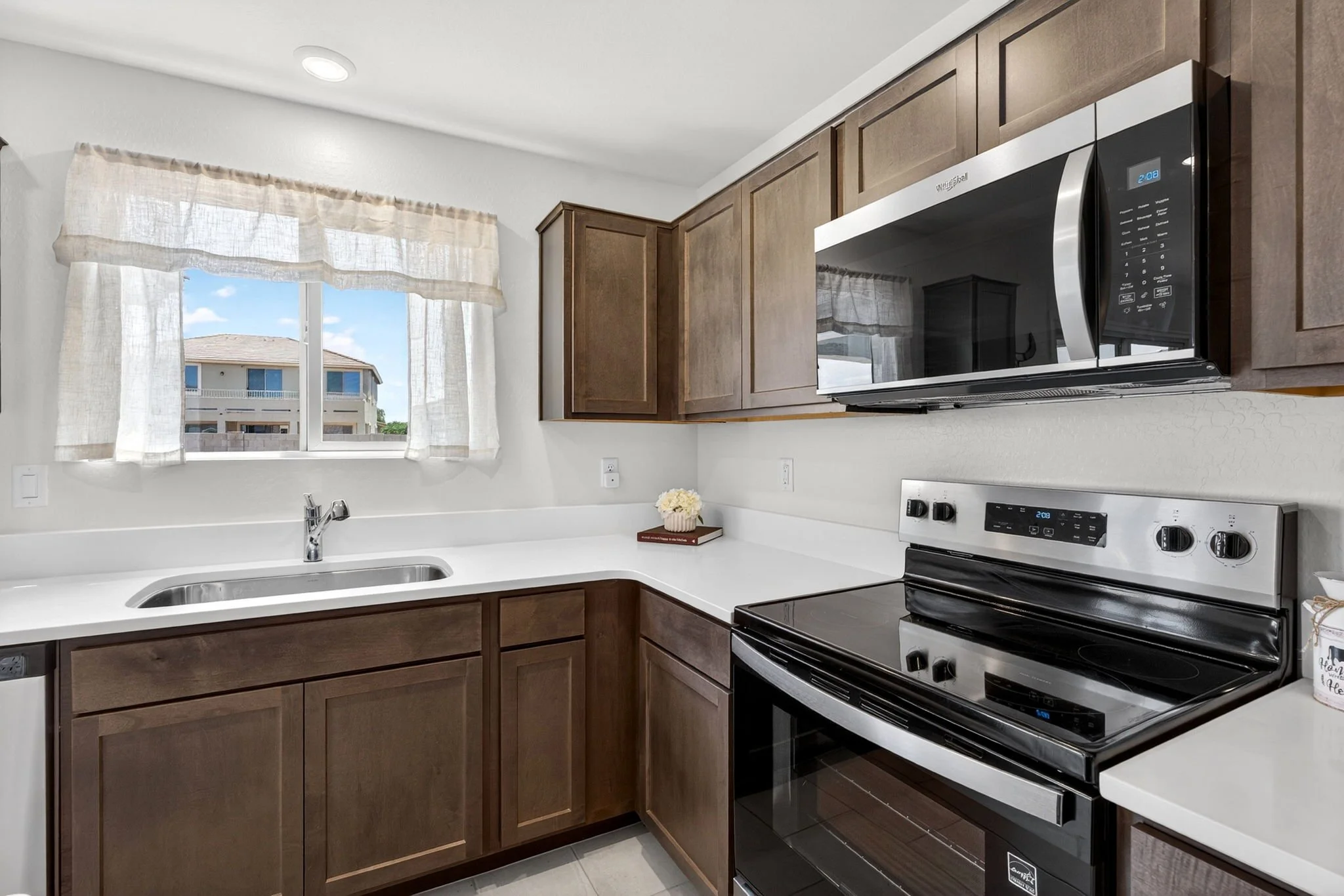 Kitchen with wooden cabinets, stainless steel microwave, electric stove, white countertop, sink, window with curtains, and view of neighboring building
