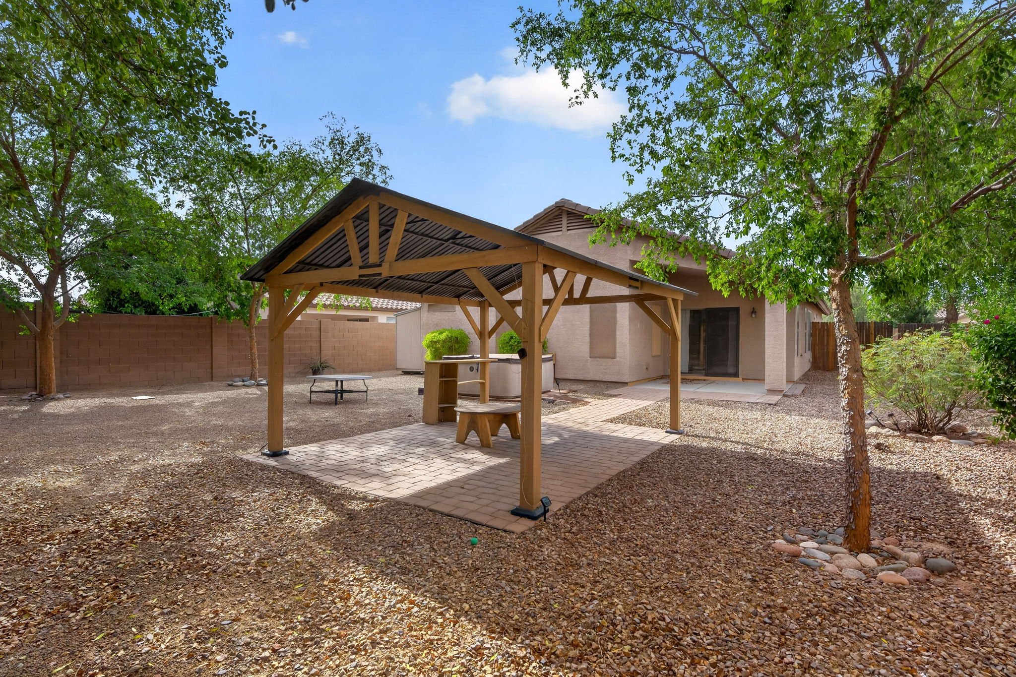 A backyard with a covered patio area, trees, a brick pathway, and a house in the background.