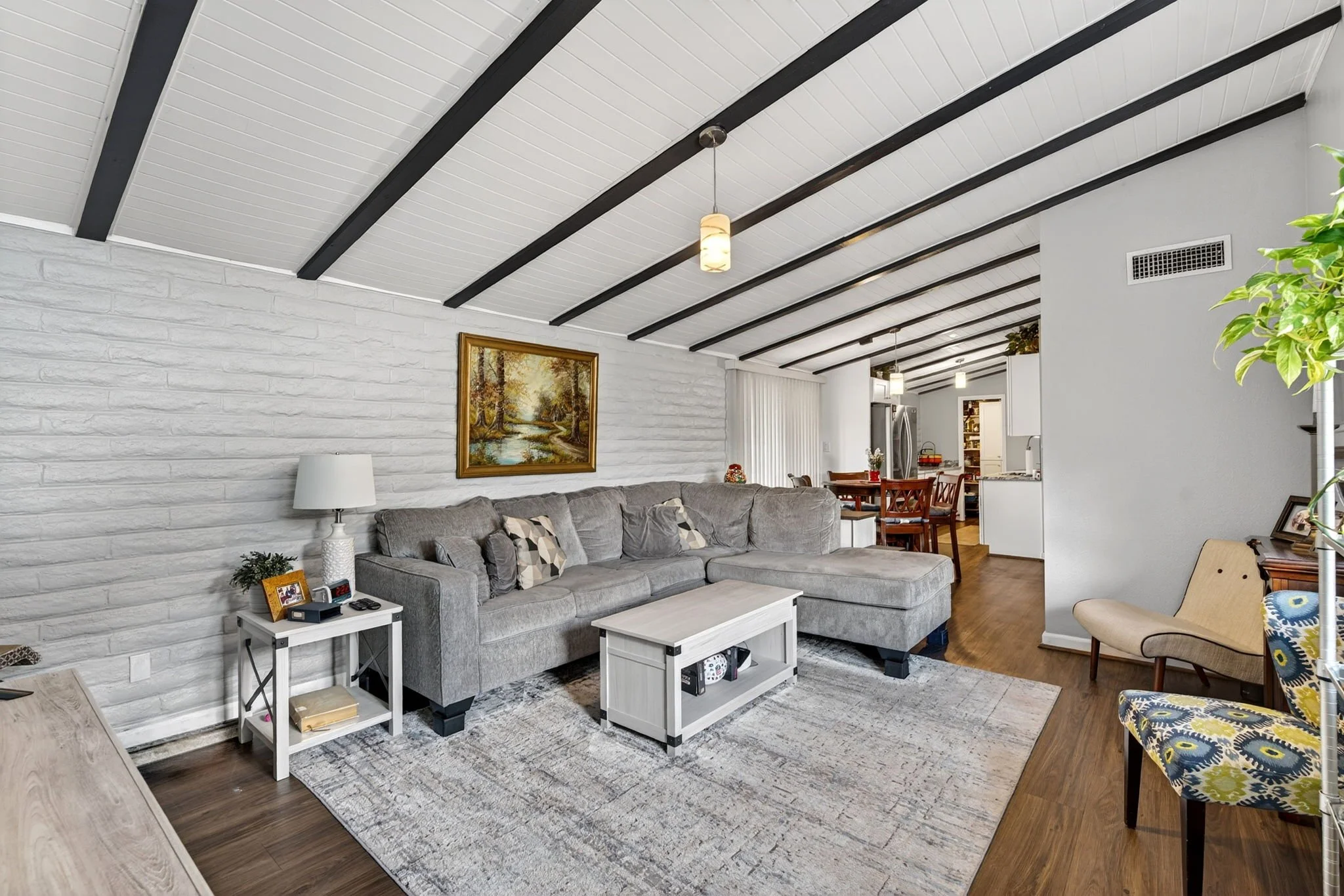 Living room with white brick wall, gray sectional sofa, artwork, white side tables, and wood flooring. Open concept with dining and kitchen area visible.