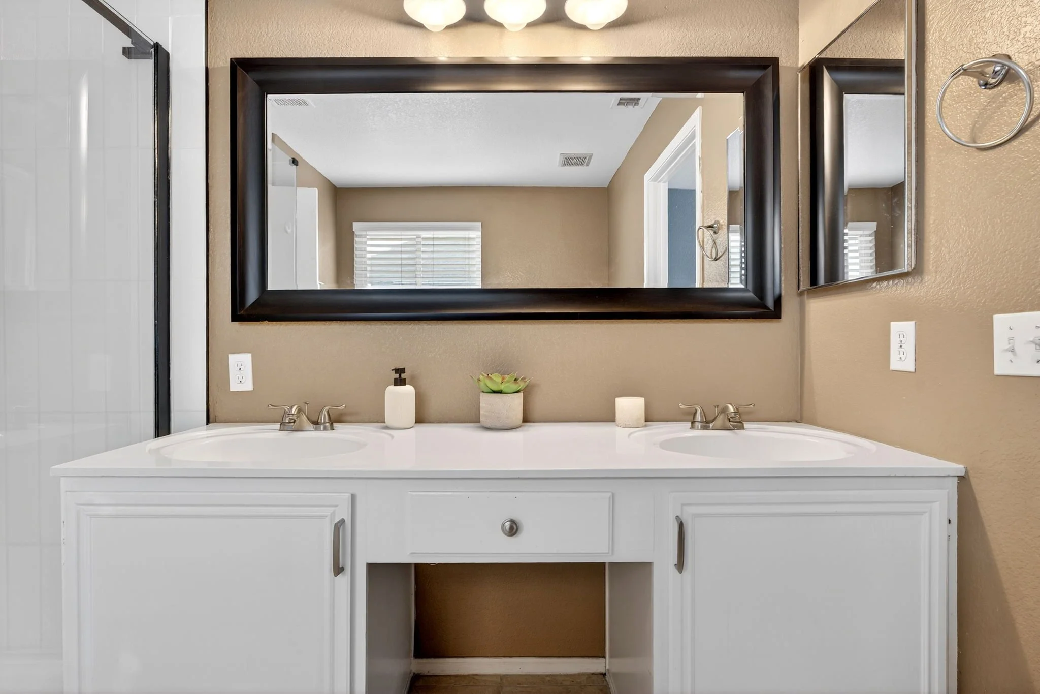 Bathroom vanity with a white cabinet, two sinks, silver faucets, and a shelf with a potted plant, two candles, a soap dispenser, and a mirror above the sinks, with a beige wall and window in the background.