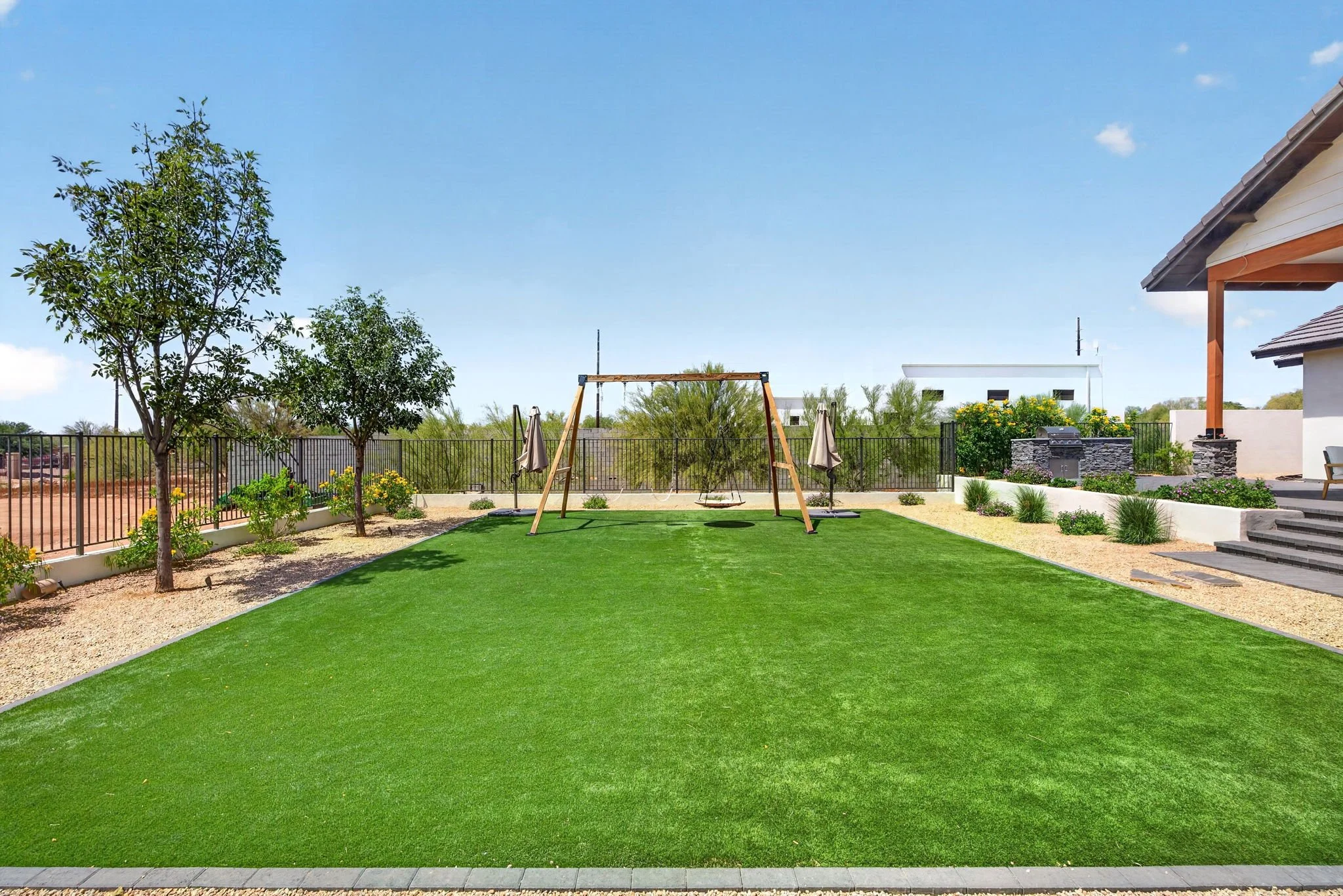 A backyard with a well-maintained grass lawn, trees, a swing set, a small vegetable garden, and a modern house with a stone chimney and outdoor steps.