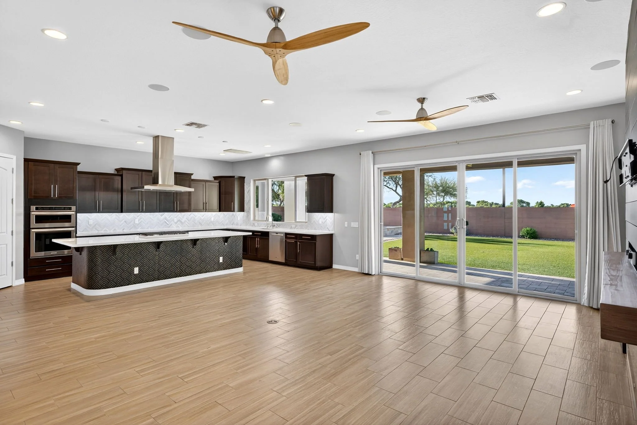 Open-concept kitchen and living area with wood flooring, dark wood cabinets, a kitchen island, ceiling fans, and sliding glass doors leading to a backyard with a lawn and trees.