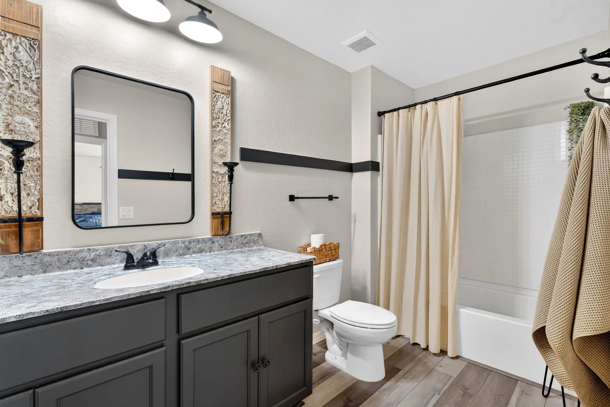 A modern bathroom with a black vanity, granite countertop, oval sink, and black fixtures. There is an oval mirror above the sink, two decorative wall panels, a black towel bar, and a shower area with cream-colored curtains. The bathroom has white wal