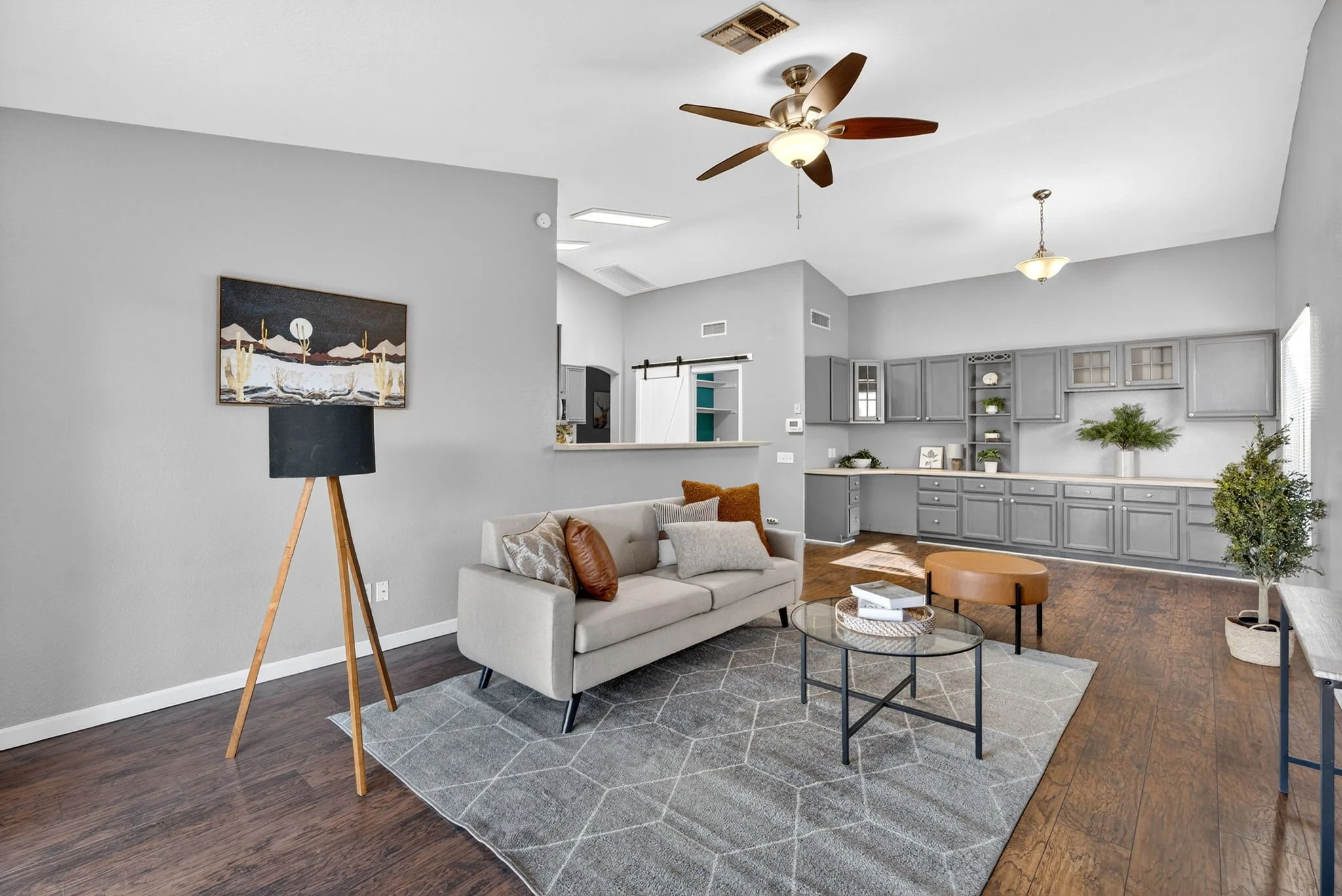 Living room with beige sofa, wooden coffee table, pendant light, ceiling fan, gray walls, a kitchen with gray cabinets, plants, and artwork, and hardwood floors.