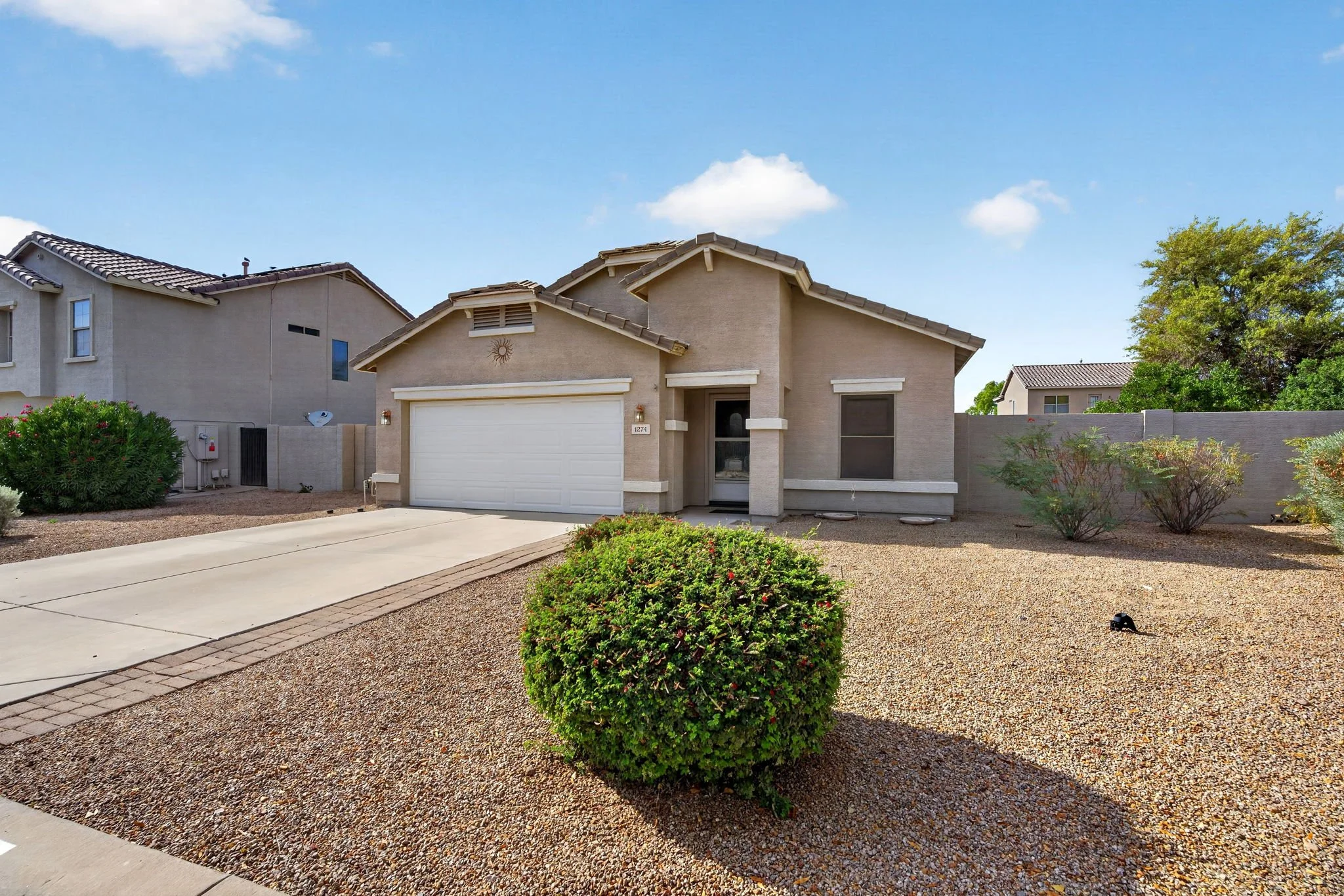 A modern beige house with a white garage door and a small front yard featuring a green shrub and gravel ground, with neighboring houses and trees in the background under a partly cloudy blue sky.