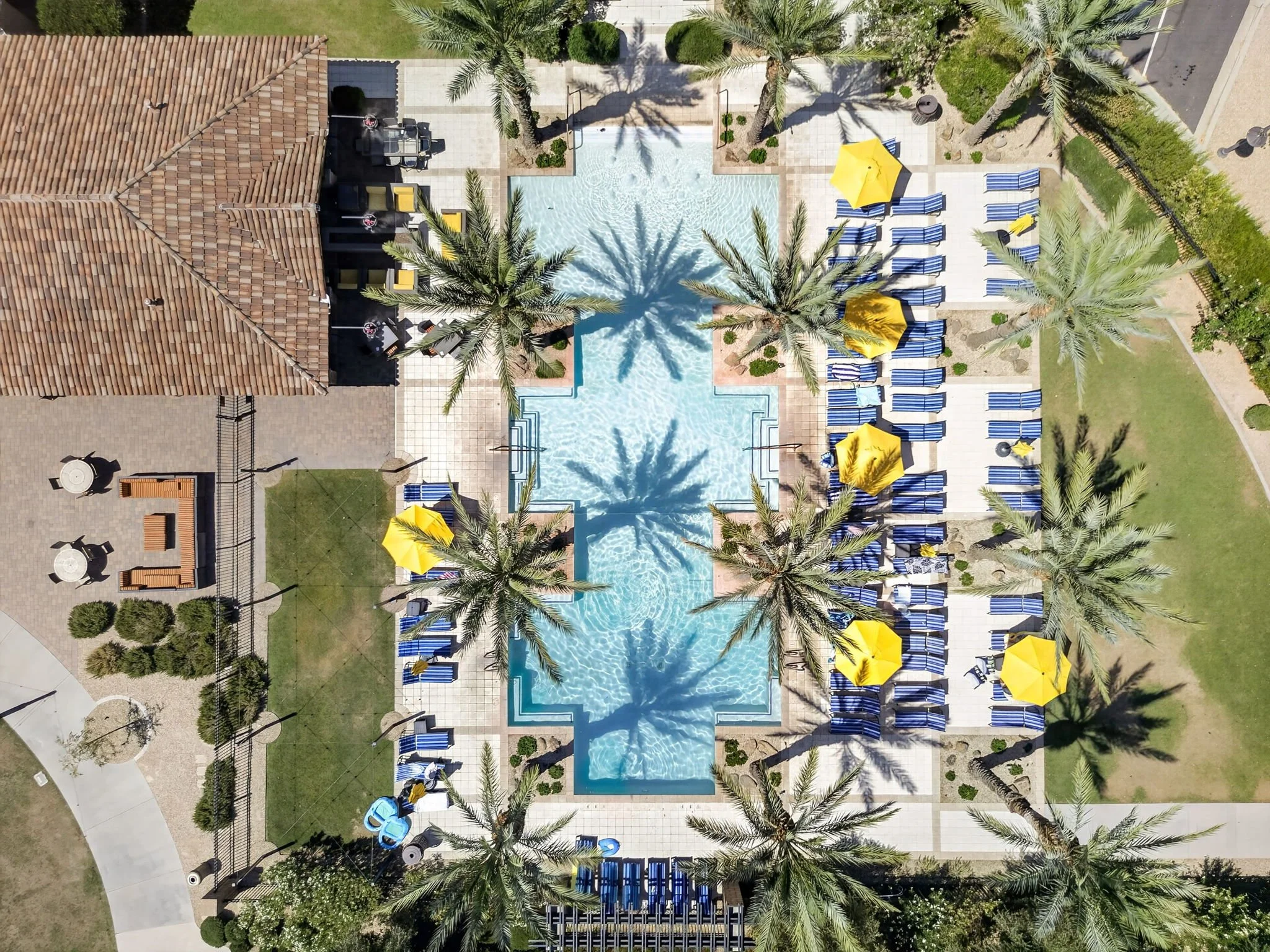 Aerial view of a swimming pool area at a resort, surrounded by lounge chairs with blue and white striped cushions and yellow umbrellas, tall palm trees, and a restroom building with a red-tiled roof.