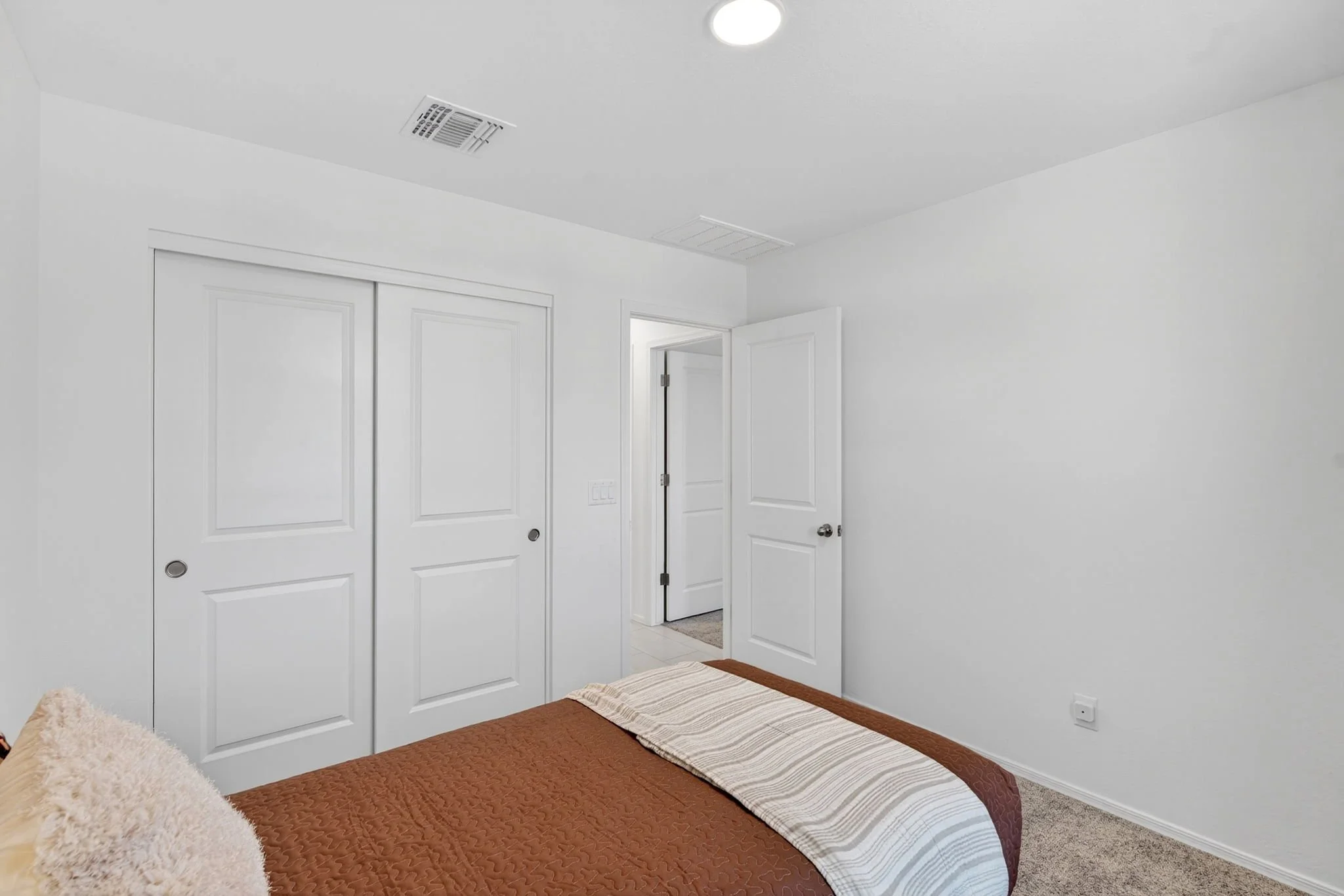A bedroom with white walls, a brown quilted bedspread, a striped pillow, and an open door leading to a hallway. There are white double closet doors and a ceiling light. The room has a beige carpet.