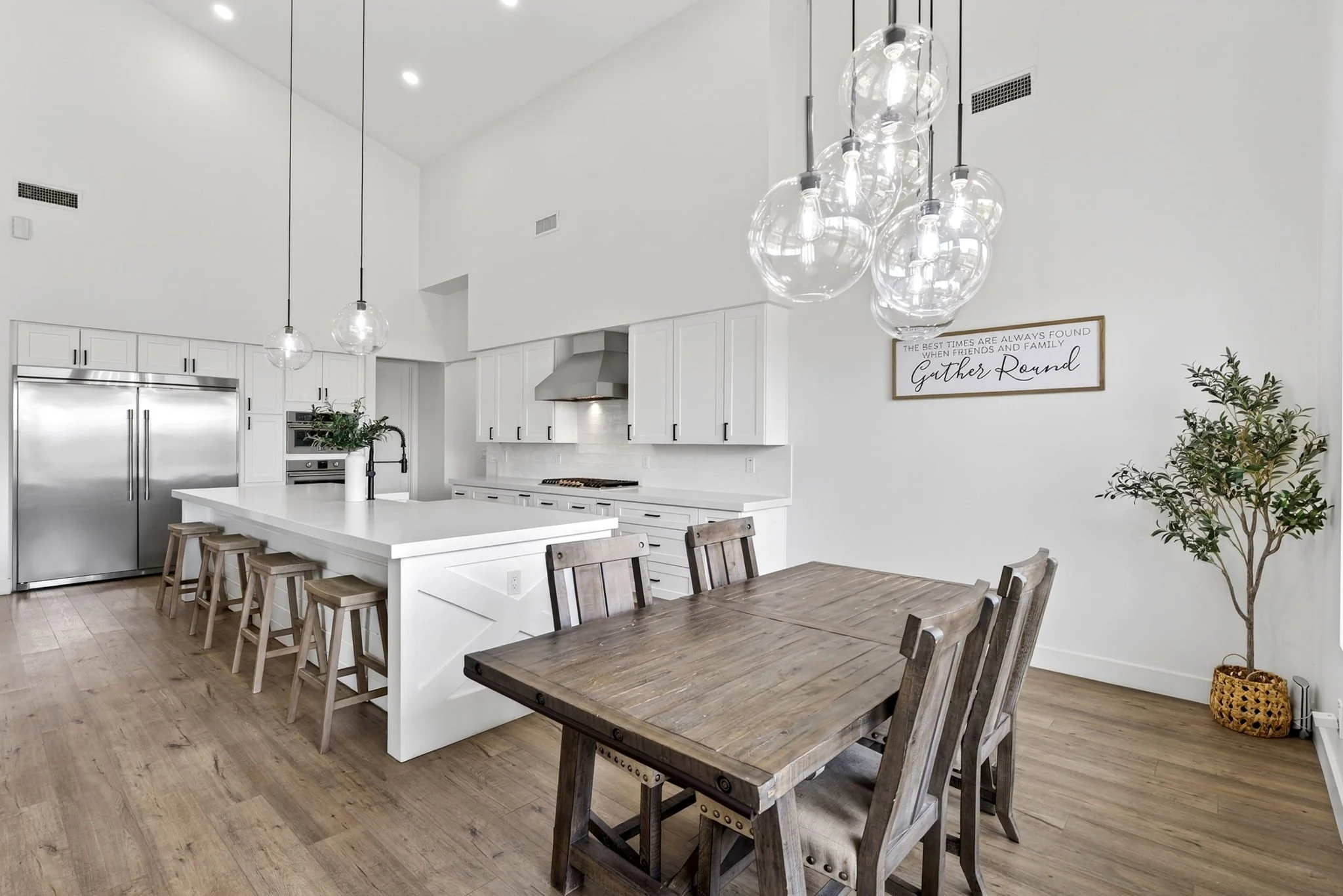 Modern kitchen with white cabinets, stainless steel appliances, and a white island with wooden stools. There is a dining table with wooden chairs, pendant lights hanging from the ceiling, and a framed quote on the wall. A potted plant and a decorativ