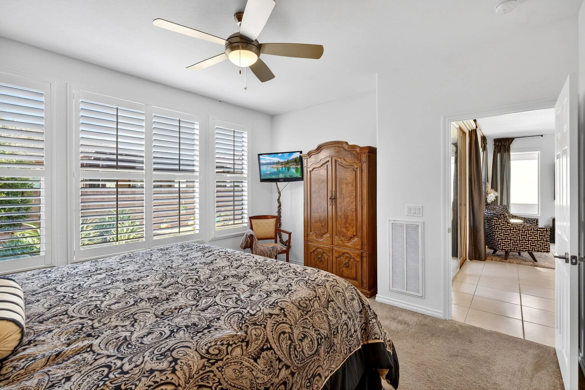 A bedroom with a bed featuring a decorative black and beige patterned bedspread. The room has large white window shutters, a wooden wardrobe, a small tv, and a chair. In the adjacent room, there is another seating area with an armchair and curtains.