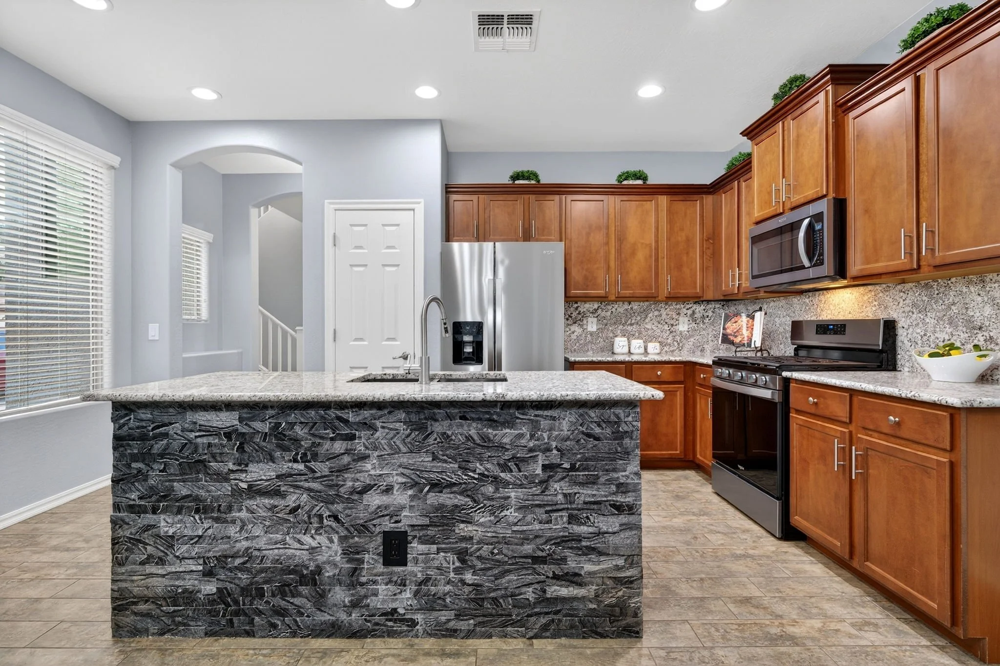 A modern kitchen features wooden cabinets, a granite countertop, a stainless steel microwave, and a stove. The kitchen island has a textured stone finish, and there are three small decorative plants on top of the upper cabinets. Large windows with wh