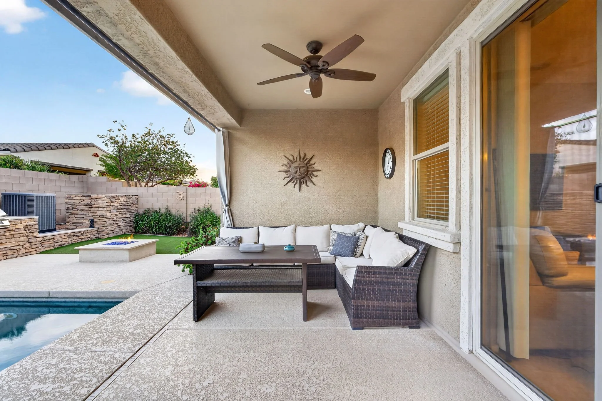 Covered backyard patio with ceiling fan, a white sectional sofa with pillows, a rectangular coffee table, a fire pit, a stone wall, and some trees and plants in the background.