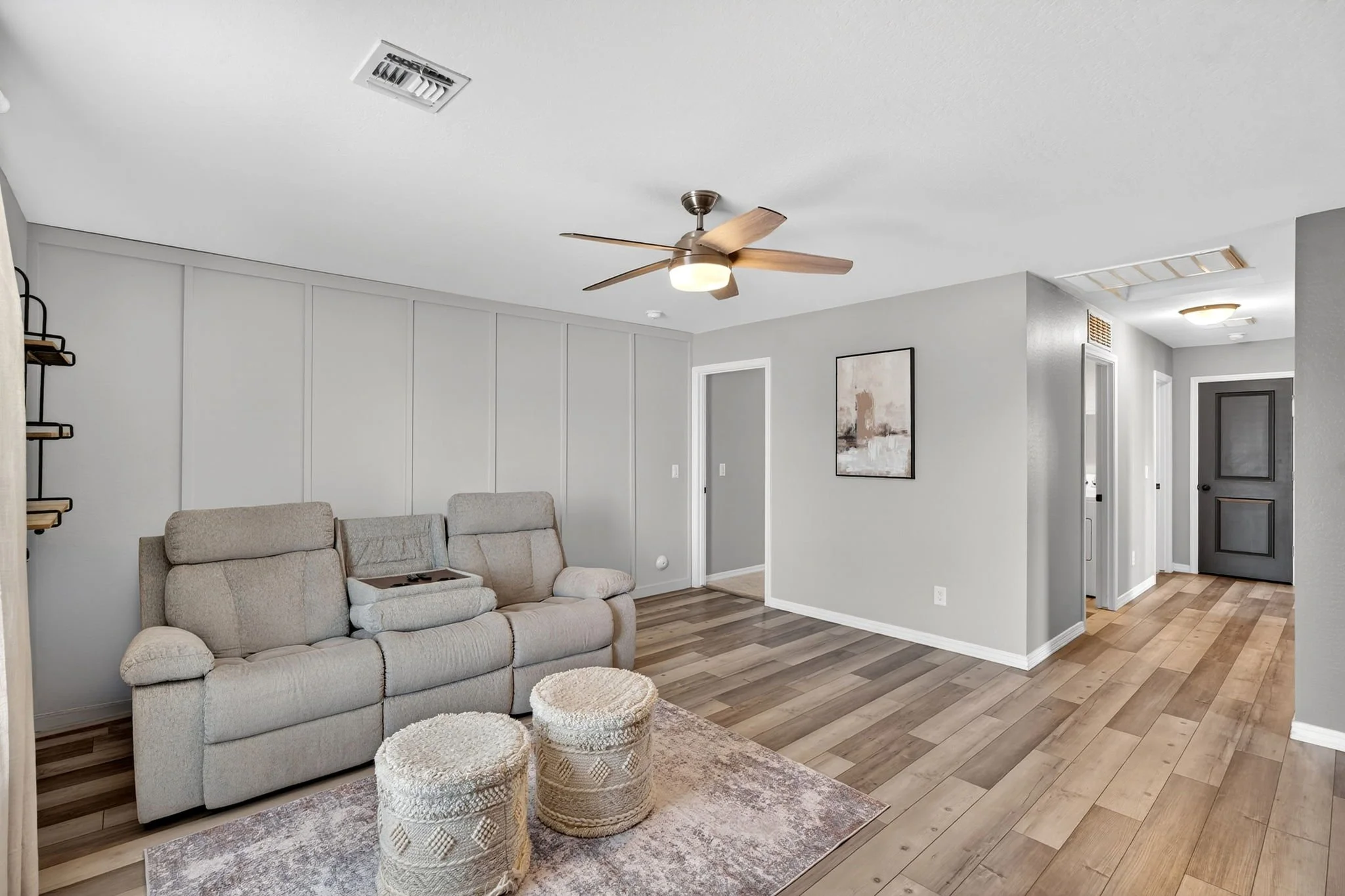 Living room with beige sofa, three woven ottomans, wall art, ceiling fan, and hallway.