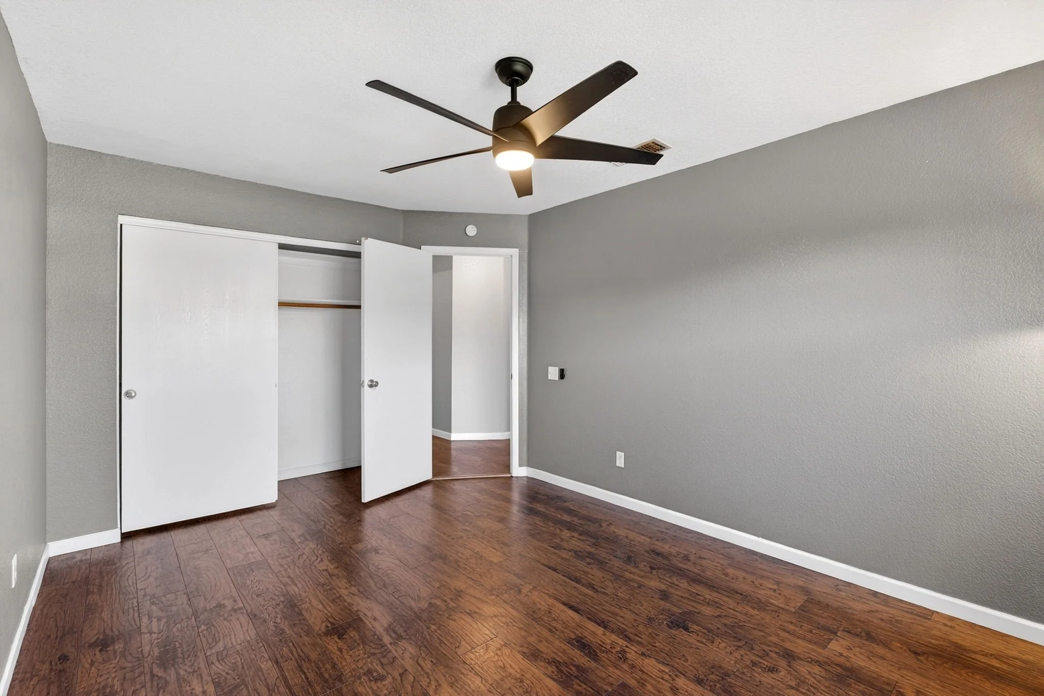 Empty bedroom with gray walls, hardwood floor, floor closet doors, and ceiling fan with light.