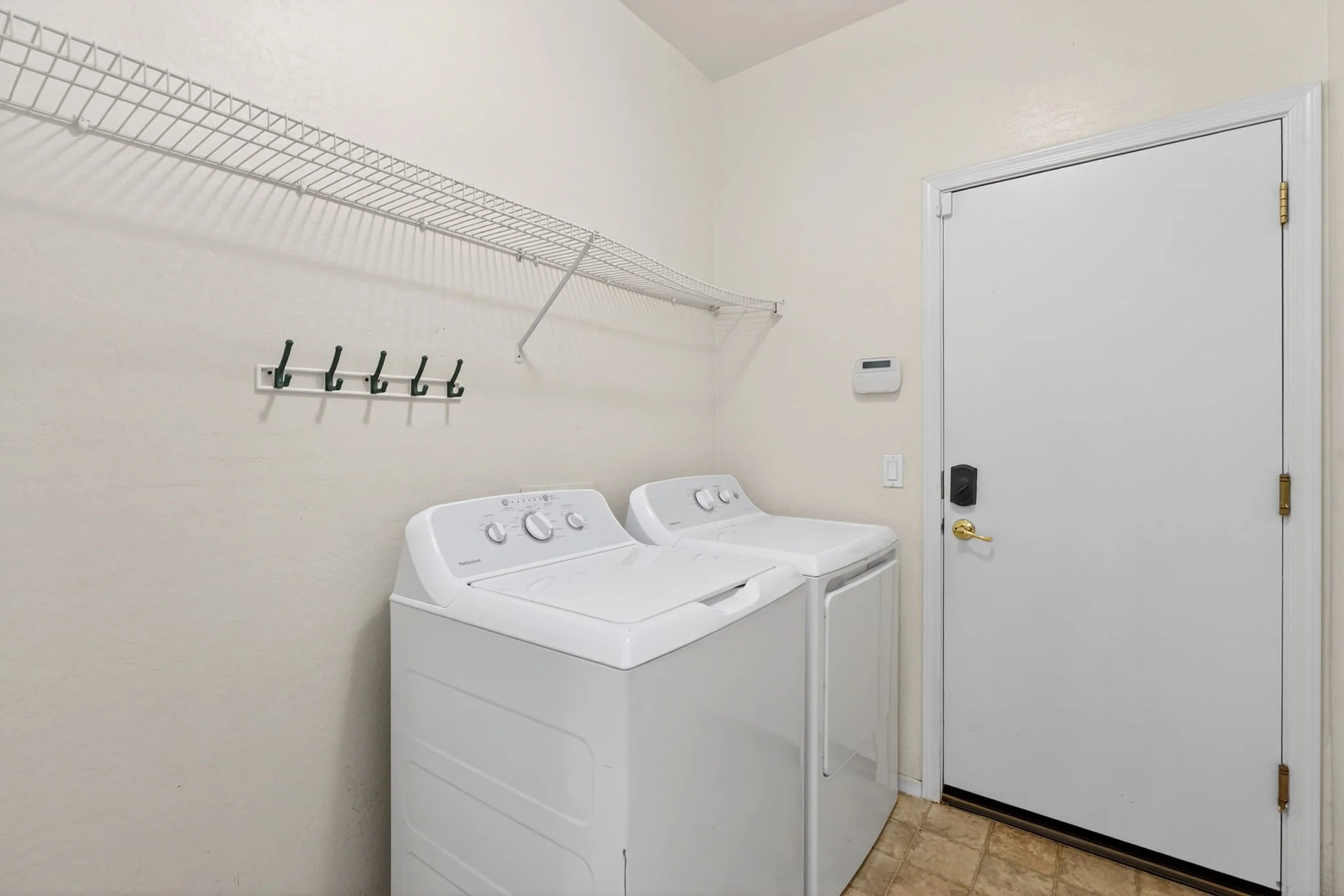 Laundry room with two white washing machines, a wire shelf above, hooks on the wall, and a door with a lock.