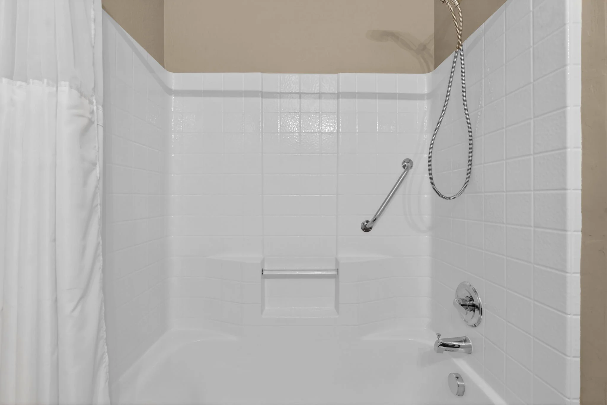 A clean, white shower area with a grab bar, a handheld shower head attached with a flexible hose, and a white curtain on the left side.