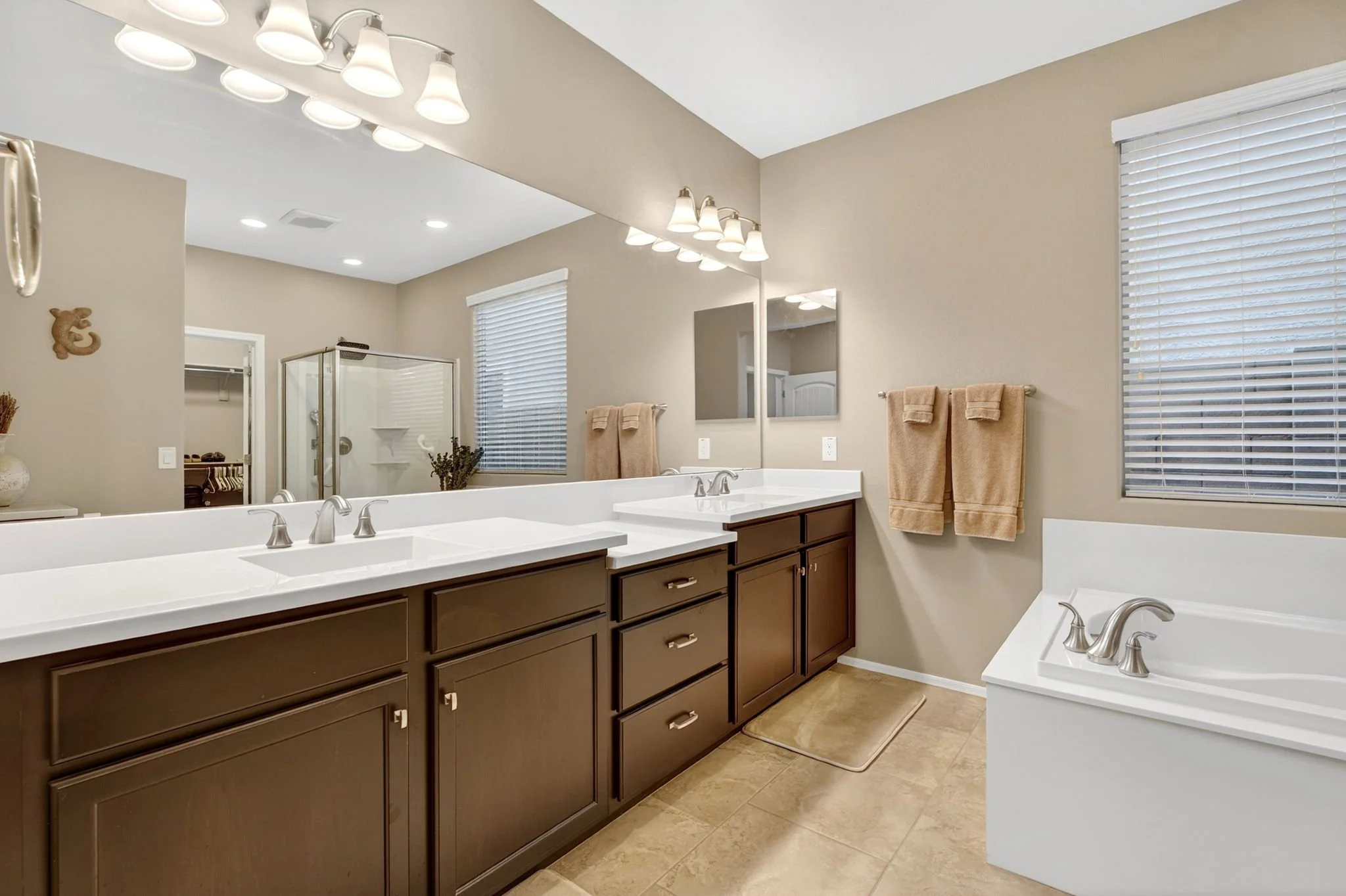 A spacious bathroom with a large mirror above a double sink vanity with dark wood cabinets. Beige towels hang on a wall, and a window with white blinds is on the side. The room is well-lit with multiple ceiling lights, and there is a separate shower 