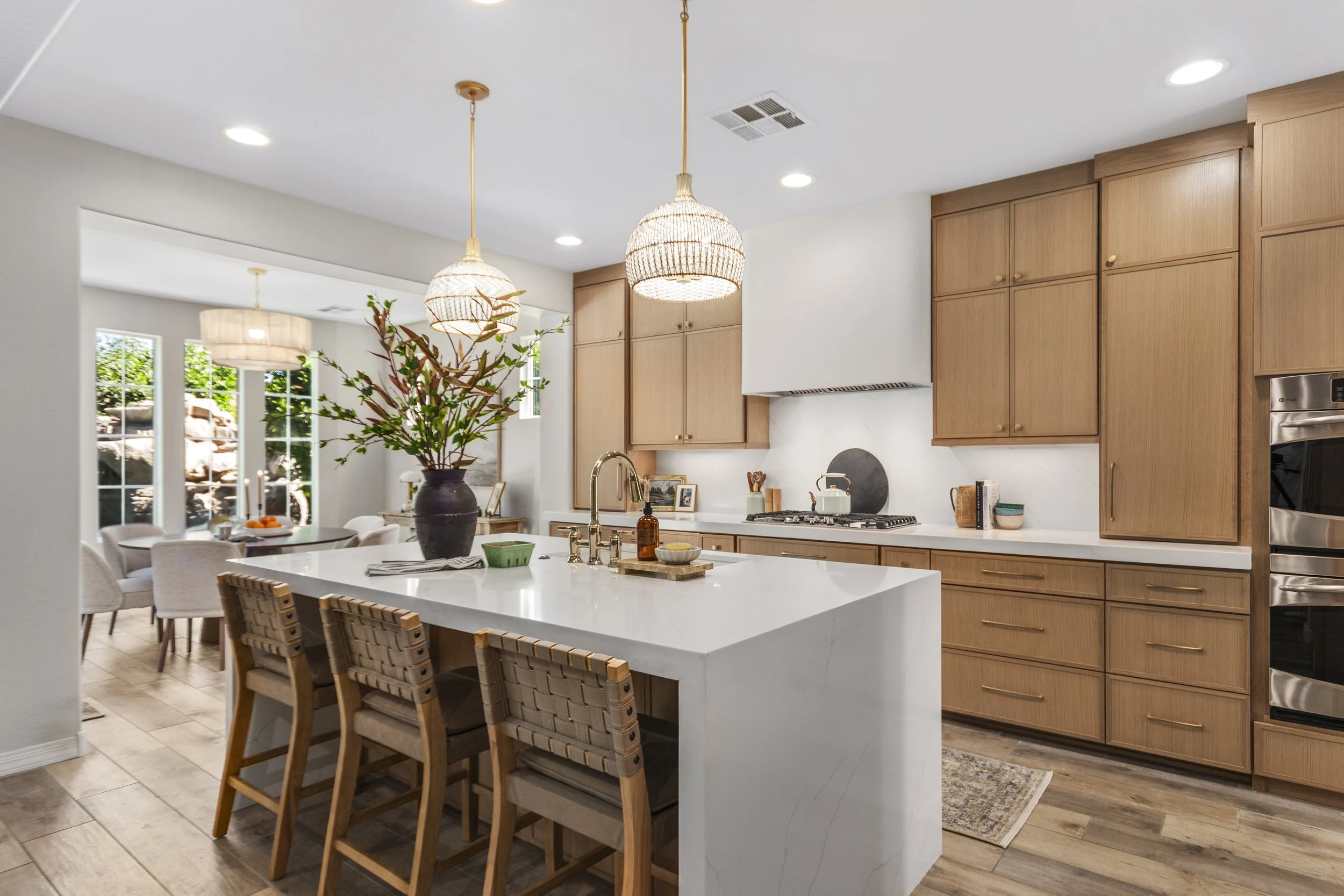 A modern kitchen with a white island counter, wooden cabinets, pendant lights, and large windows in the background letting in natural light.