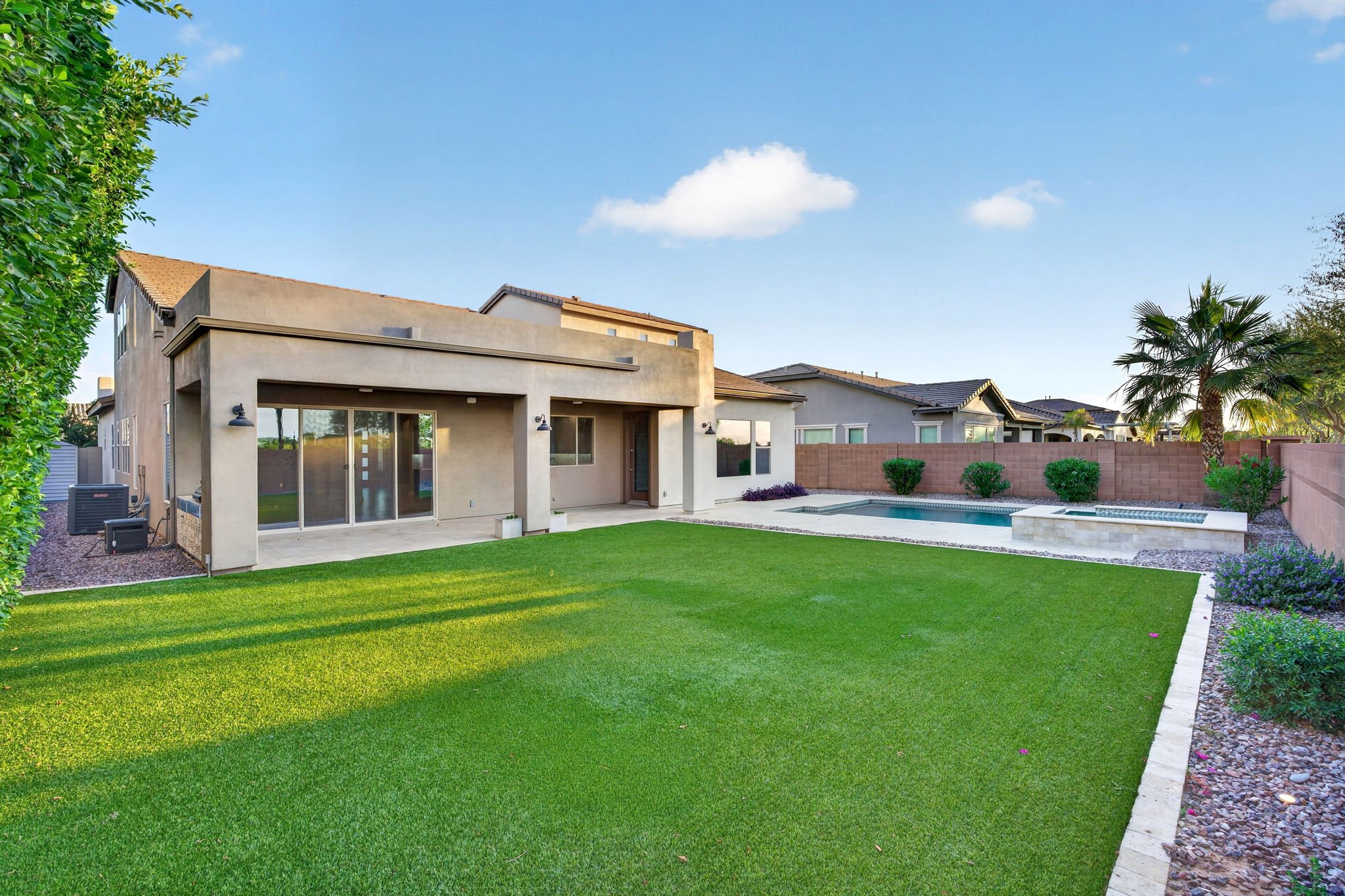 The image depicts the backyard of a modern house with a neatly maintained green lawn, a swimming pool, and various small bushes. The house has large sliding glass doors, and the backyard is enclosed by a brown brick wall, with neighboring houses visi