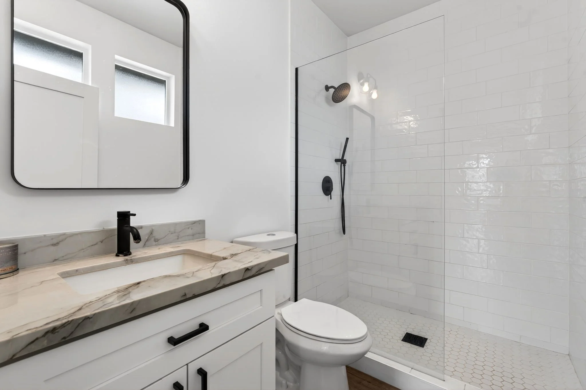 Modern bathroom with white walls, a marble vanity with black fixtures, a large mirror, a white toilet, and a shower area with white subway tiles and black fixtures.