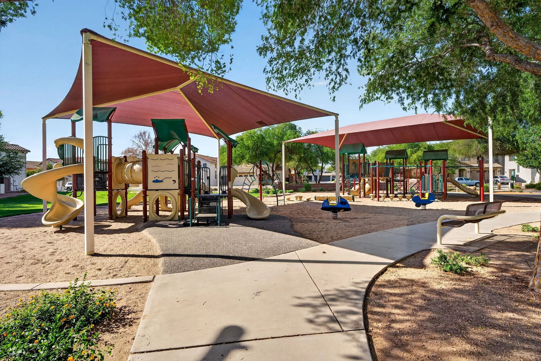 Children's playground with slides, swings, and climbing structures under shaded canopies in a park, with trees and residential buildings in the background.
