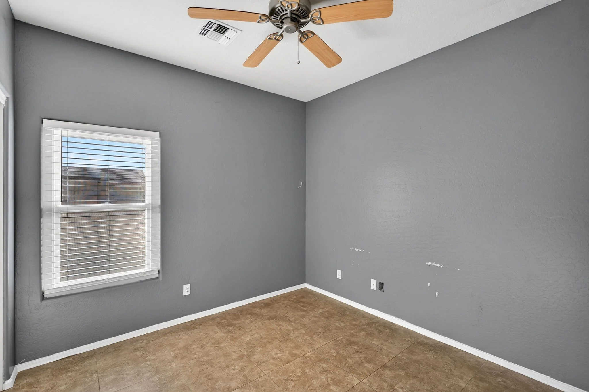 Empty room with gray walls, white trim, a window with white blinds, a ceiling fan with wooden blades, and a brown tile floor.
