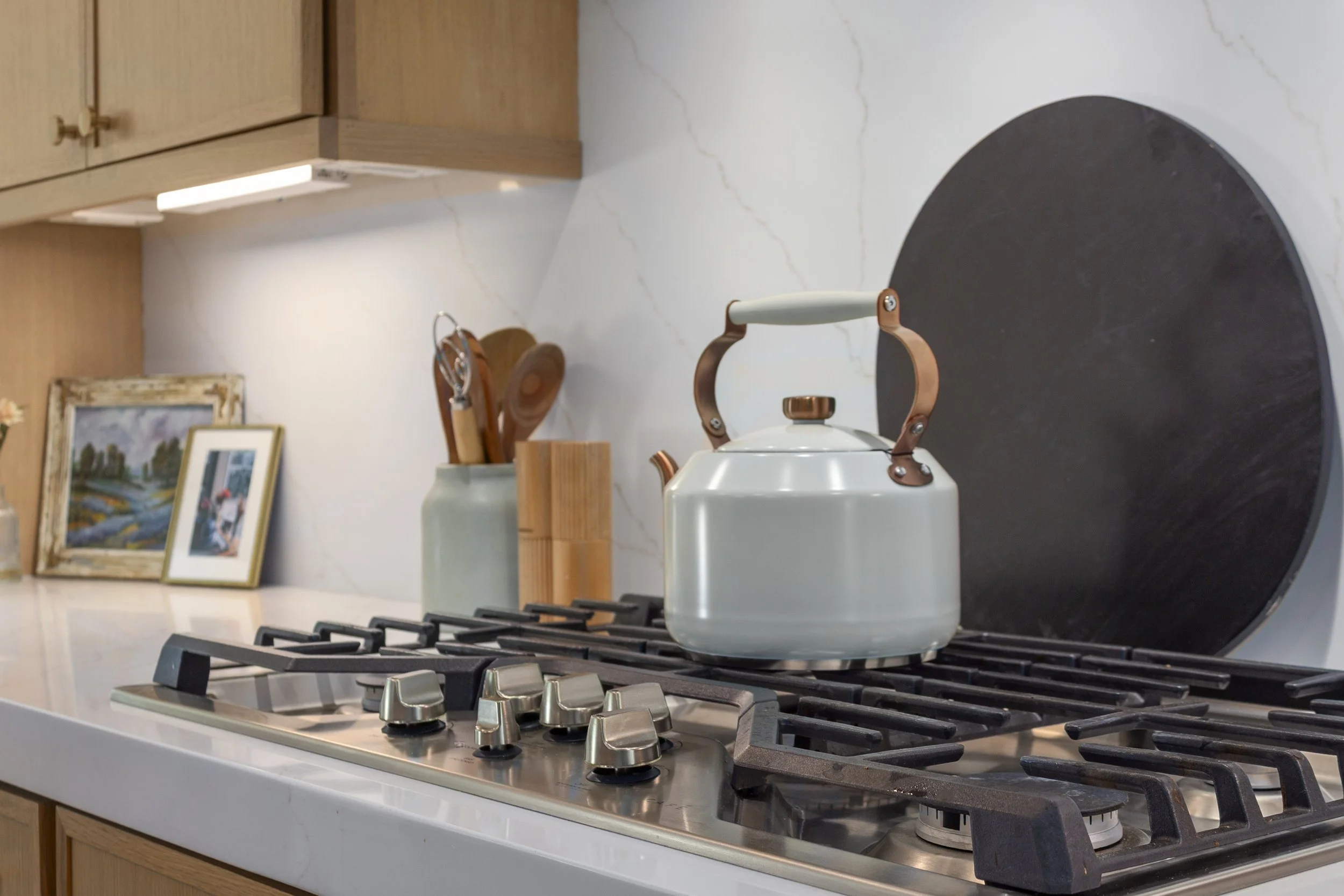 White kettle with copper accents on a gas stove with black grates, background includes wooden cabinet, framed pictures, and kitchen utensils.