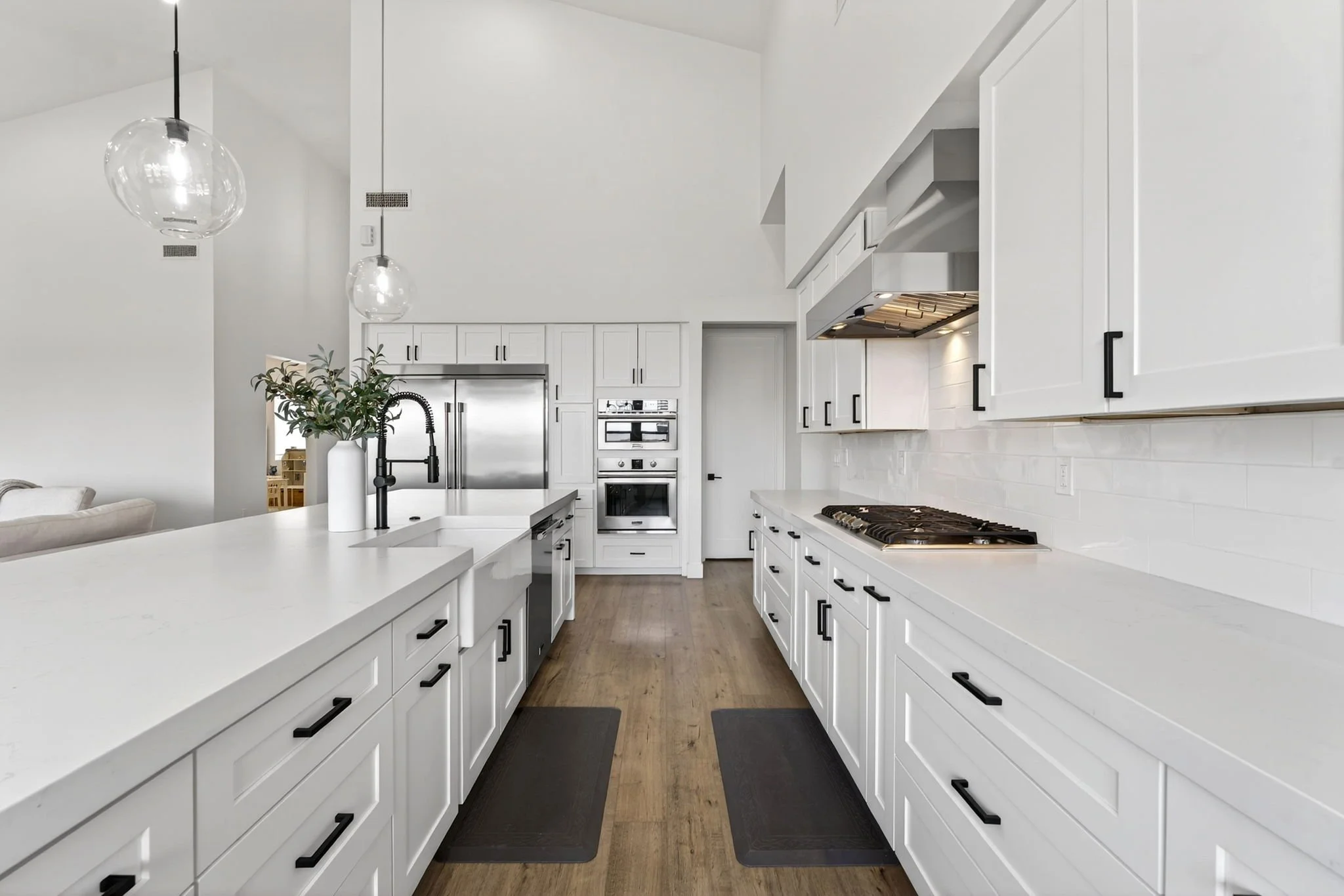 Modern white kitchen with island, black handles, stainless steel appliances, and pendant lights.