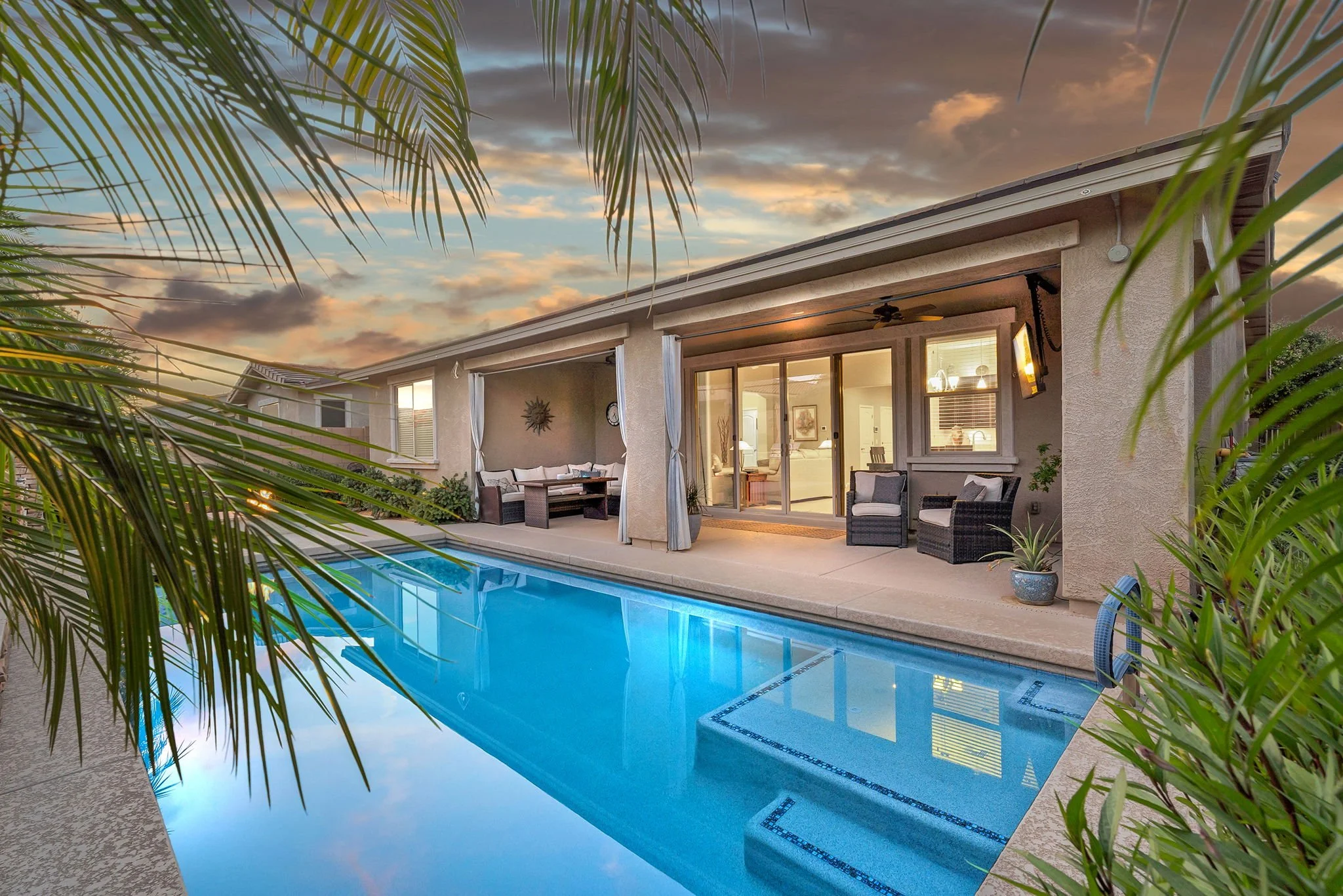 Backyard patio with a swimming pool, outdoor furniture, and a house with large sliding glass doors at sunset, surrounded by palm trees.