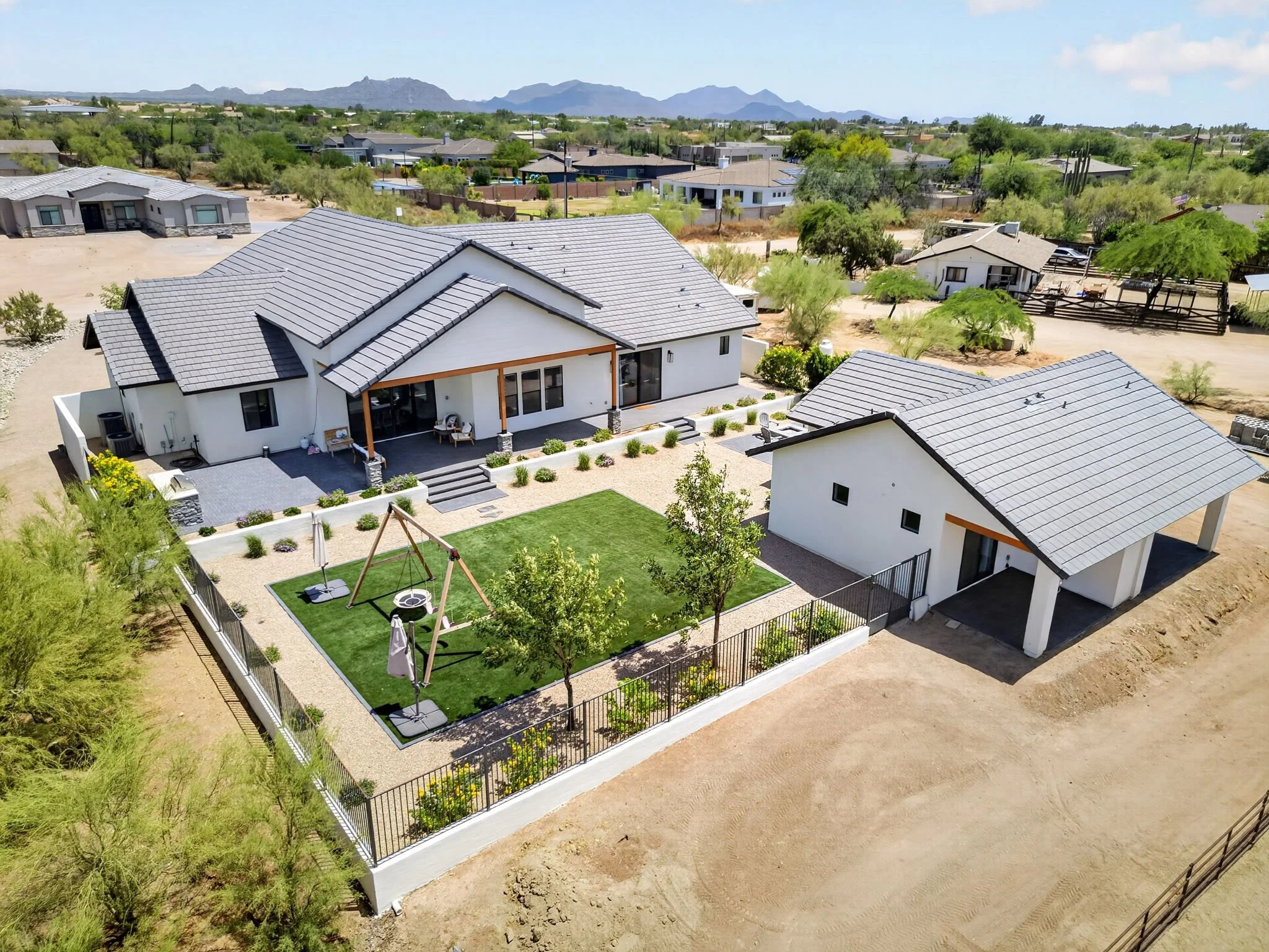 Aerial view of a modern house with a backyard, playground, and a grassy area, in a suburban neighborhood with desert landscape and mountains in the background.