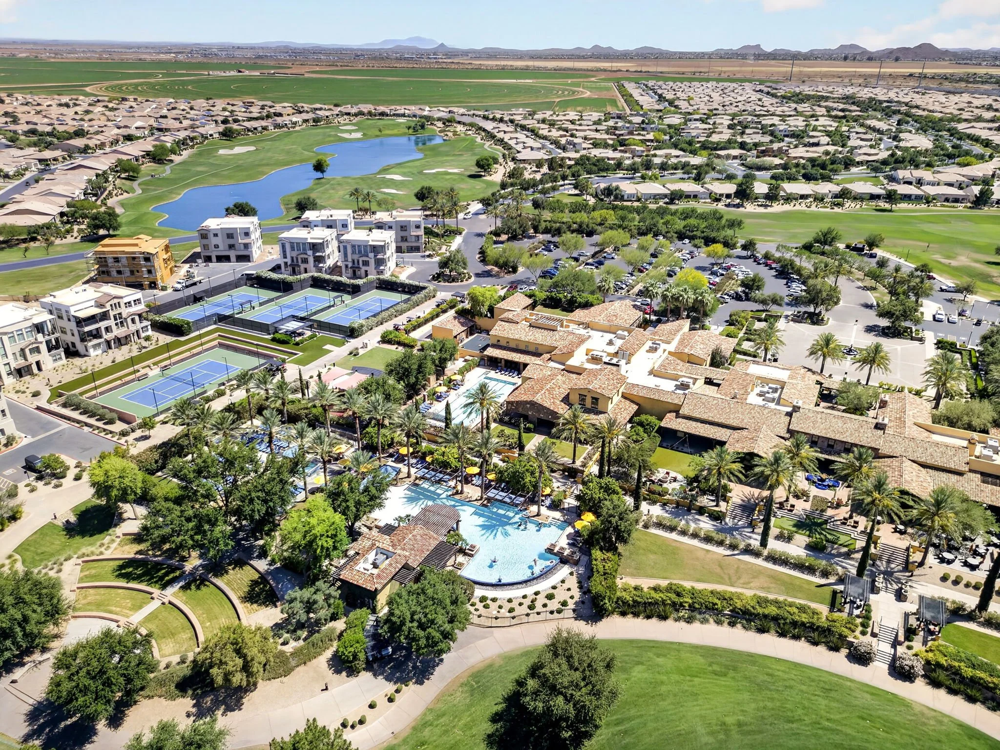 Aerial view of a residential community with a golf course featuring green fairways, a small lake, tennis courts, and a swimming pool, surrounded by desert landscape and distant mountains under a clear sky.