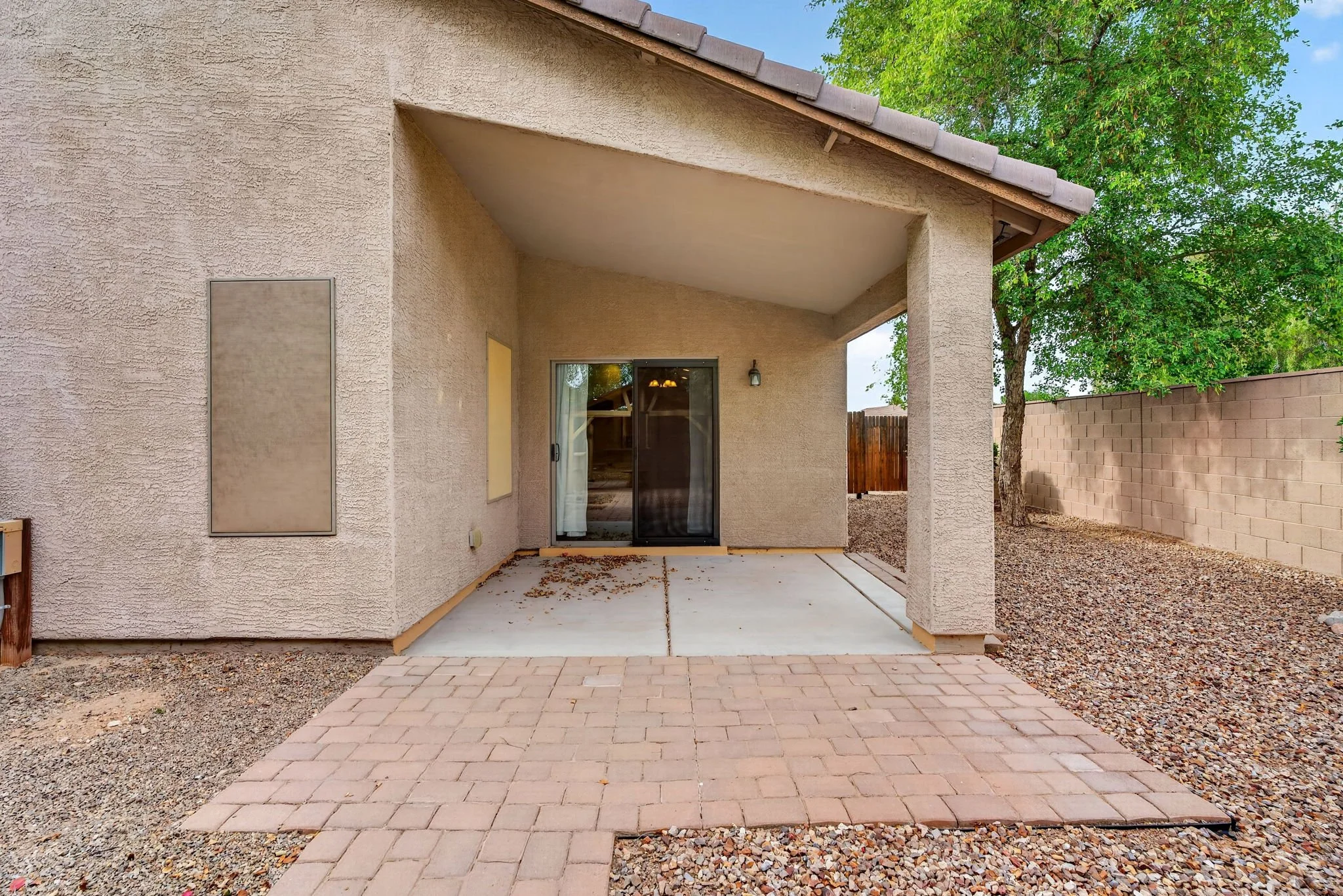 Backyard patio area with paving stones, beige stucco walls, sliding glass door, and a small roofed section. There is a tree, a wooden privacy fence, and a brick wall in the background.