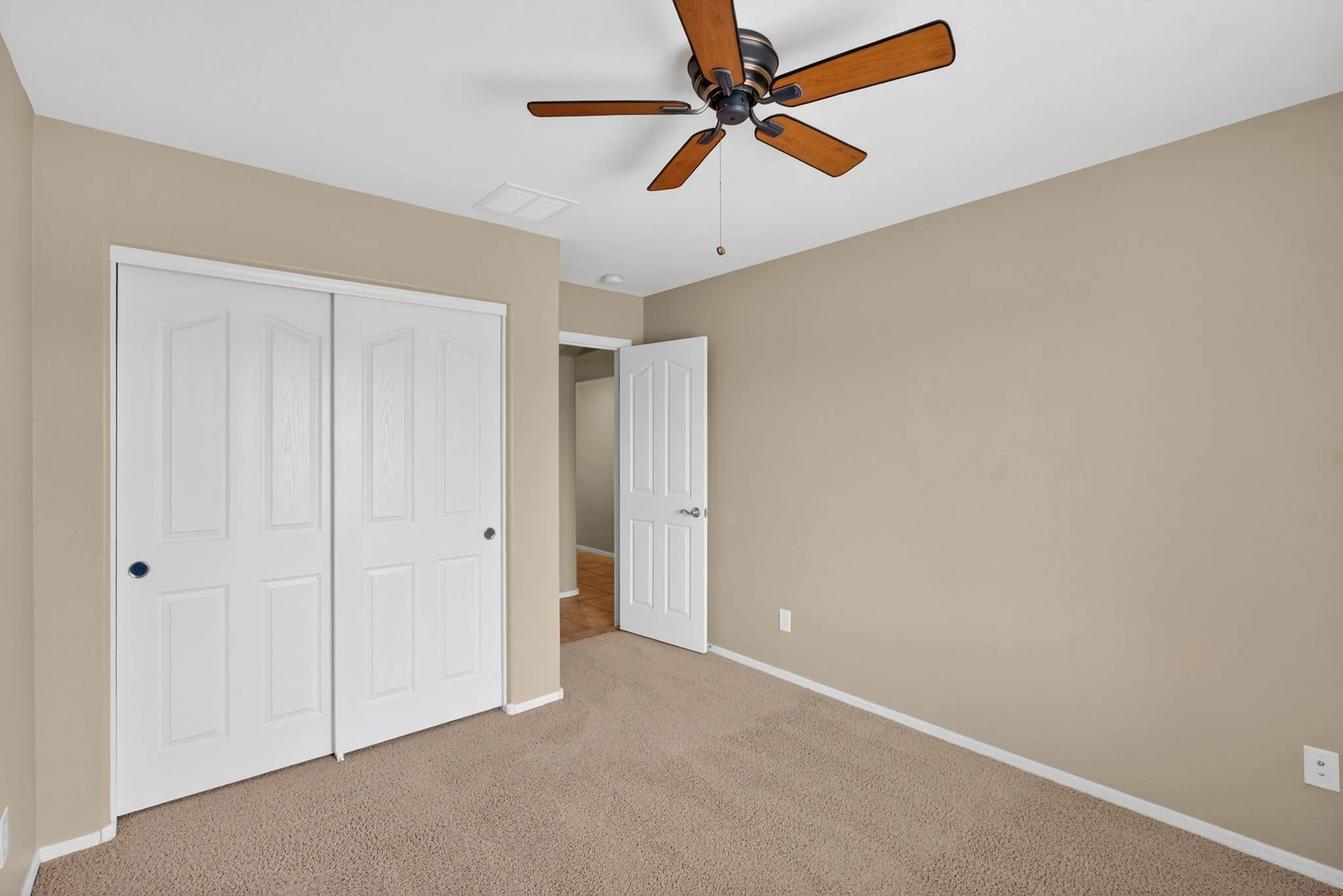 Empty room with beige walls, white double closet doors, beige carpet, and a ceiling fan with wooden blades.