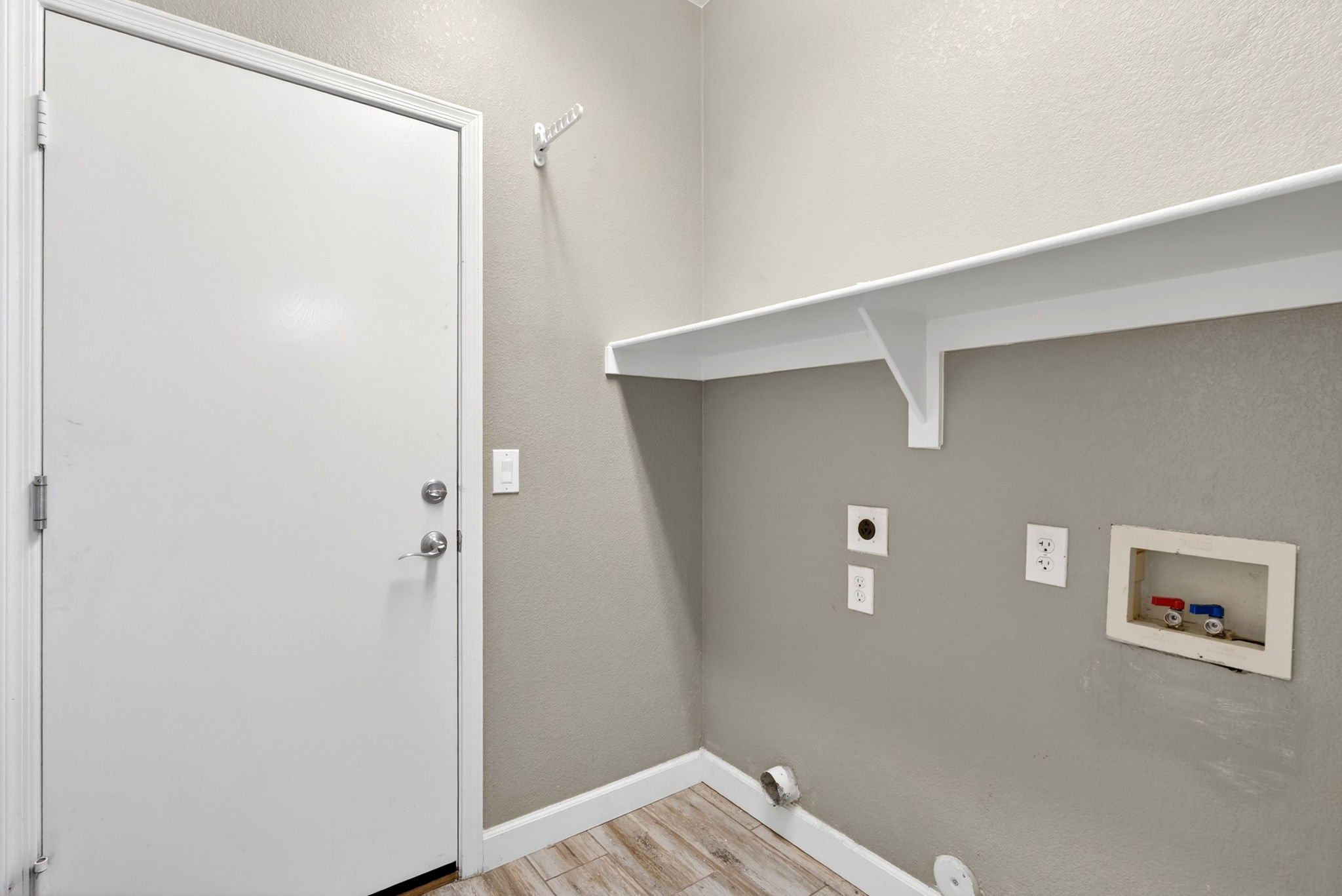 An unfinished utility room with a white door on the left, gray walls, a white shelf mounted on the wall, and exposed electrical outlets with red and blue connectors. The floor is wood-like laminate.