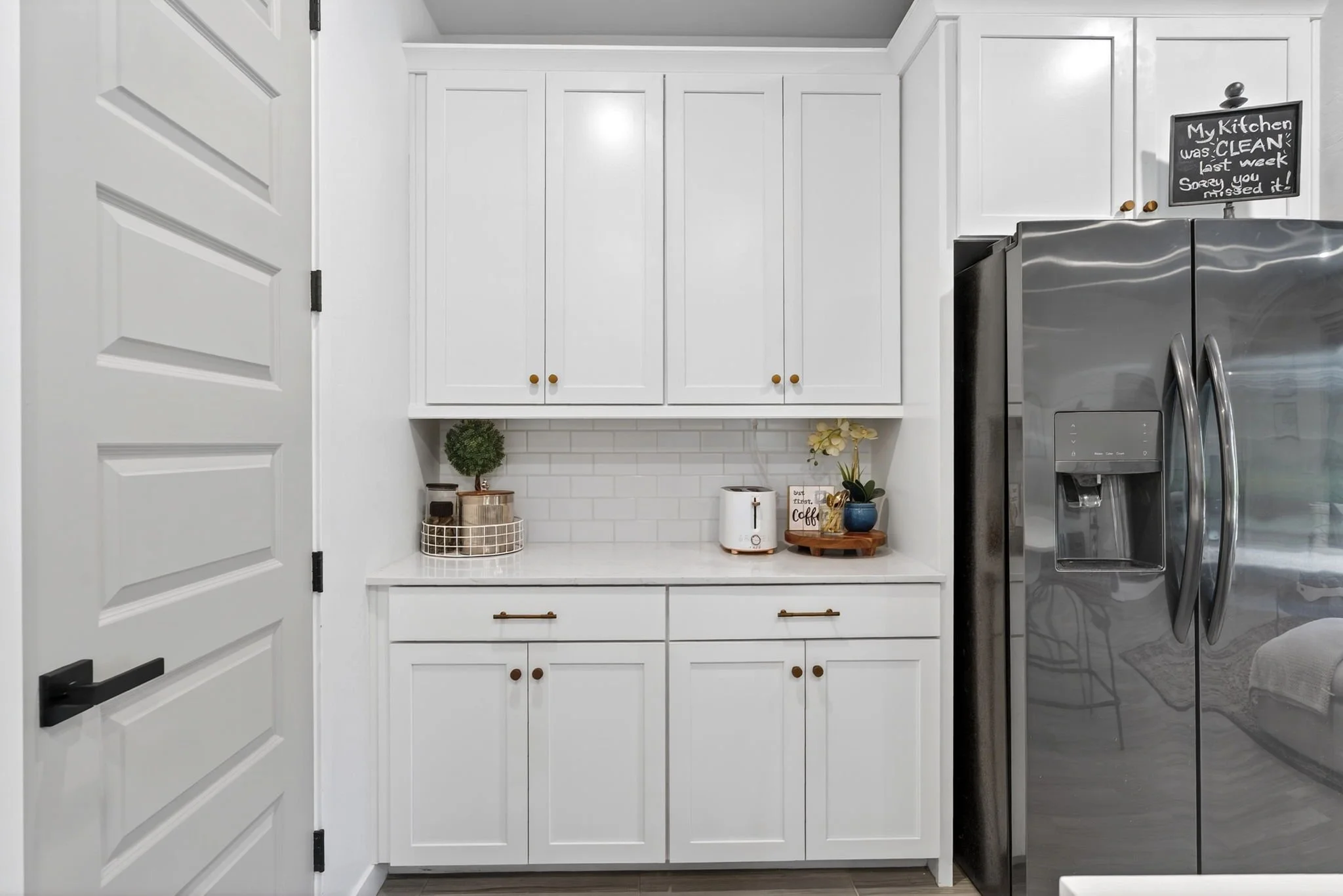 Modern white kitchen with white cabinets, stainless steel refrigerator, and decorative items on the countertop, including a small potted plant, a laundry paper towel roll, and a coffee-themed sign. A chalkboard sign on the wall reads, "My Kitchen was